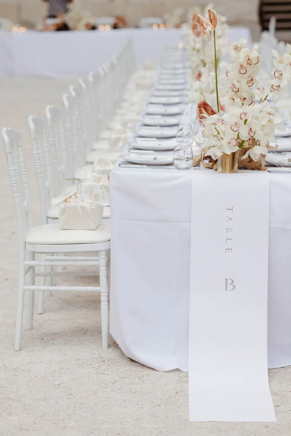 Table élégante décorée avec de grandes compositions florales d'orchidées blanches et roses dans un vase en bois, nappes blanches, verres, assiettes et couverts, invitées à un événement en plein air.