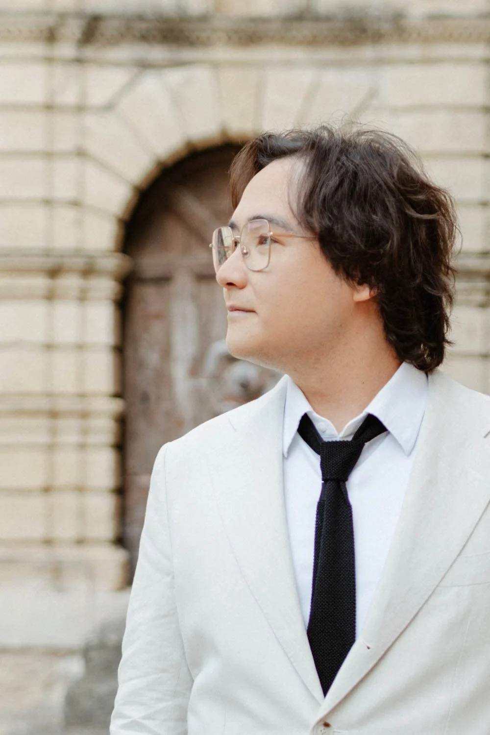 Jeune homme portant des lunettes, habillé d'un costume blanc avec une chemise blanche et une cravate noire, se tenant devant un bâtiment en pierre