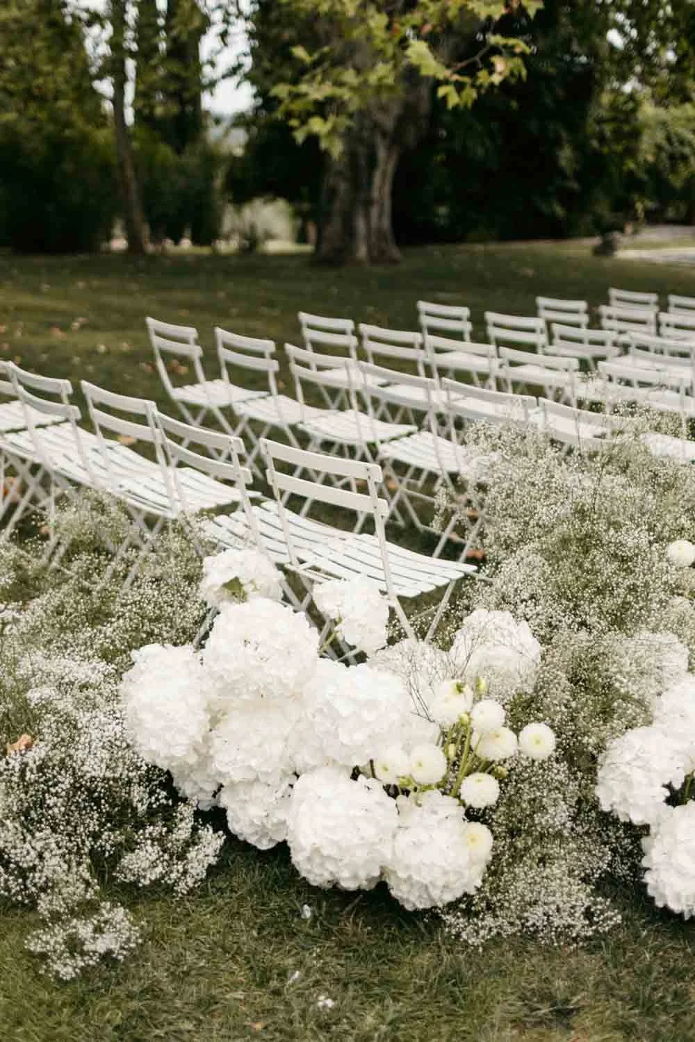 Chaises blanches disposées en rangée sur une pelouse, entourées de fleurs blanches, avec des arbres en arrière-plan, pour une cérémonie en plein air.