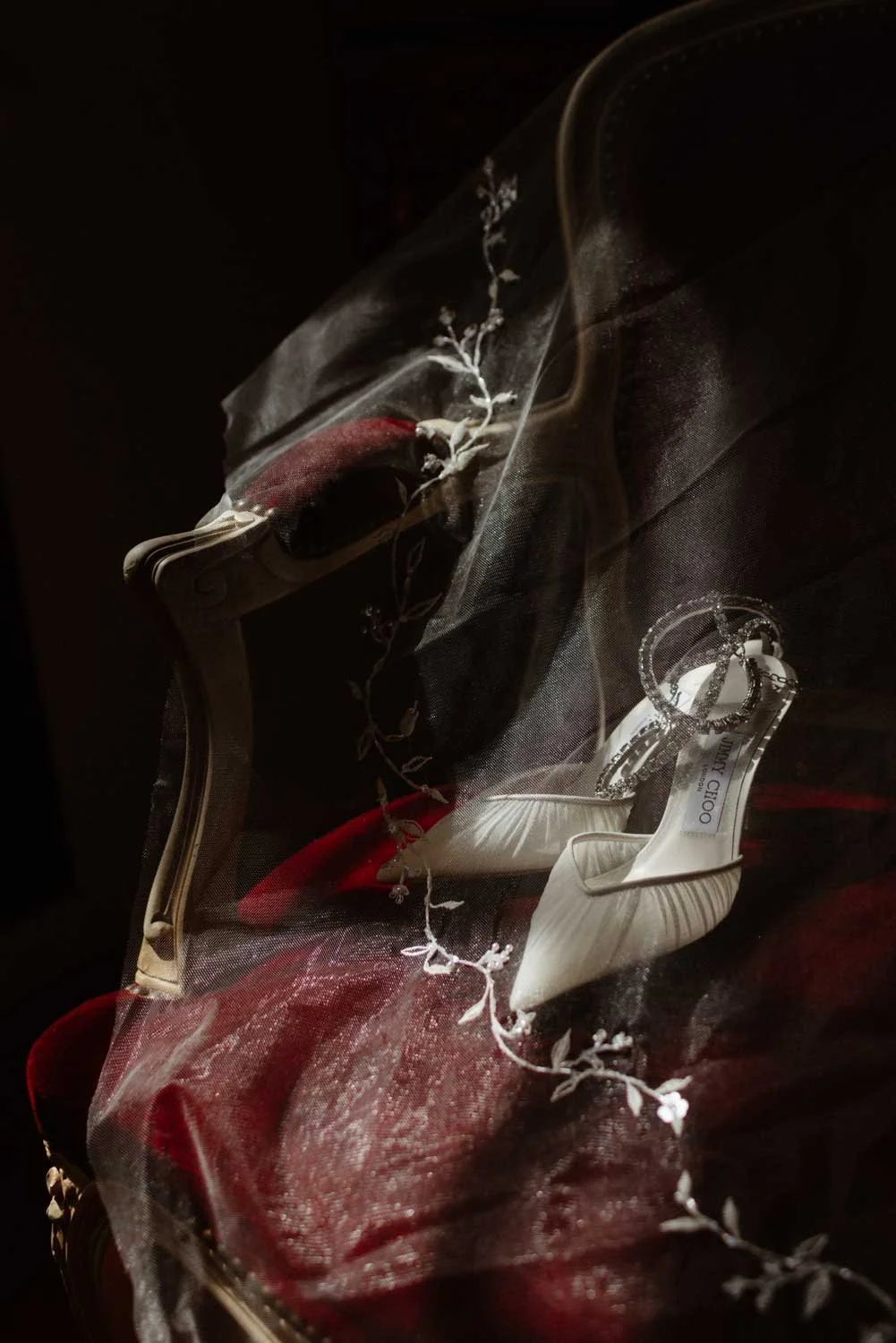 Une paire de chaussures à talons blancs avec des broderies, sous une vitre, accompagnée de décorations florales blanches et d'une pièce de bijouterie.