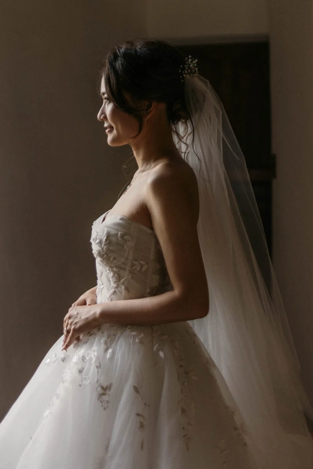 Une femme en robe de mariée, avec un voile, souriant en regardant à gauche, dans une lumière tamisée.