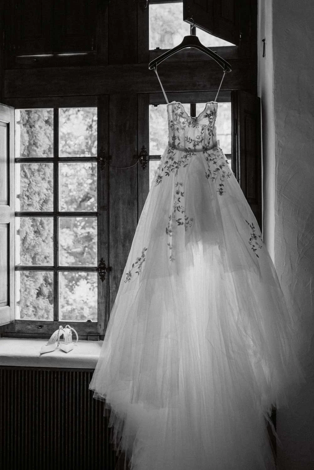 Robe de mariée suspendue devant une fenêtre en bois, avec des chaussures blanches posées sur un rebord.