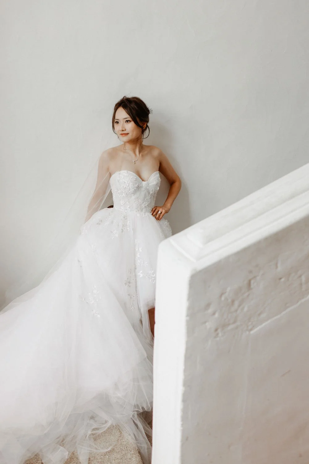 Jeune femme en robe de mariée blanche, debout près d'un mur blanc, regardant latéralement, avec une pose élégante et un sourire subtil.