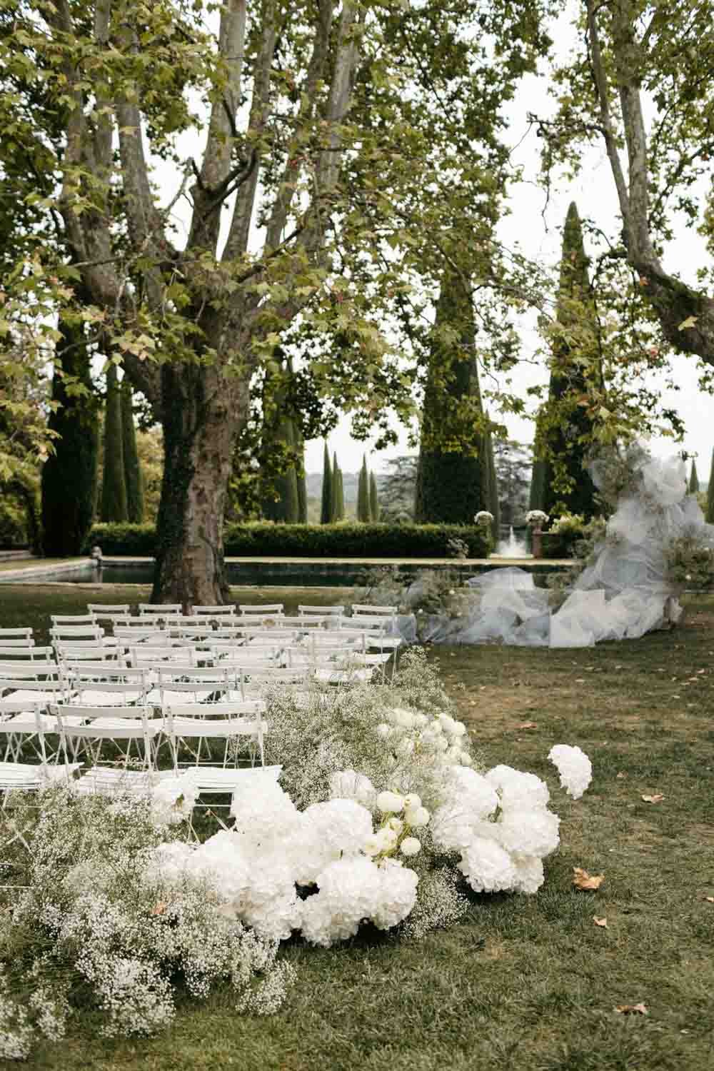 Décoration de mariage en extérieur avec des chaises blanches, des fleurs blanches et un arbre grand, dans un jardin ou un parc, avec une fontaine en arrière-plan.
