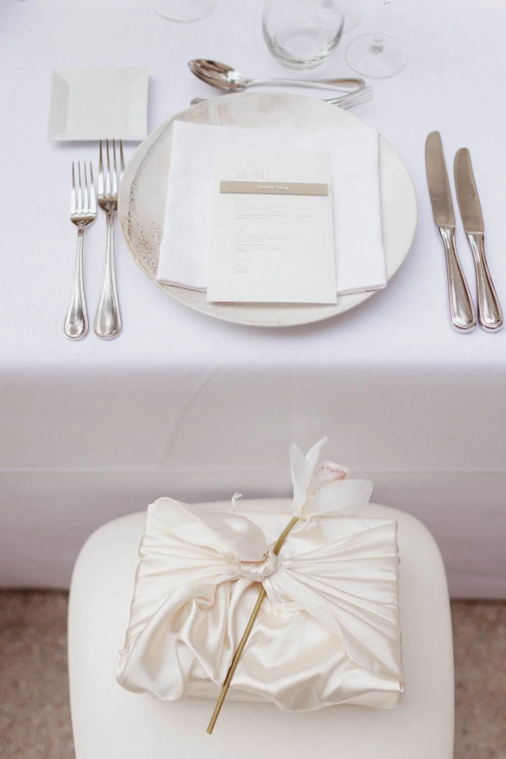 Table élégante avec une assiette, des couverts en argent, un verre à vin, un menu, et une nappe blanche. Sur une chaise en face, un paquet cadeau beige avec une orchidée blanche.