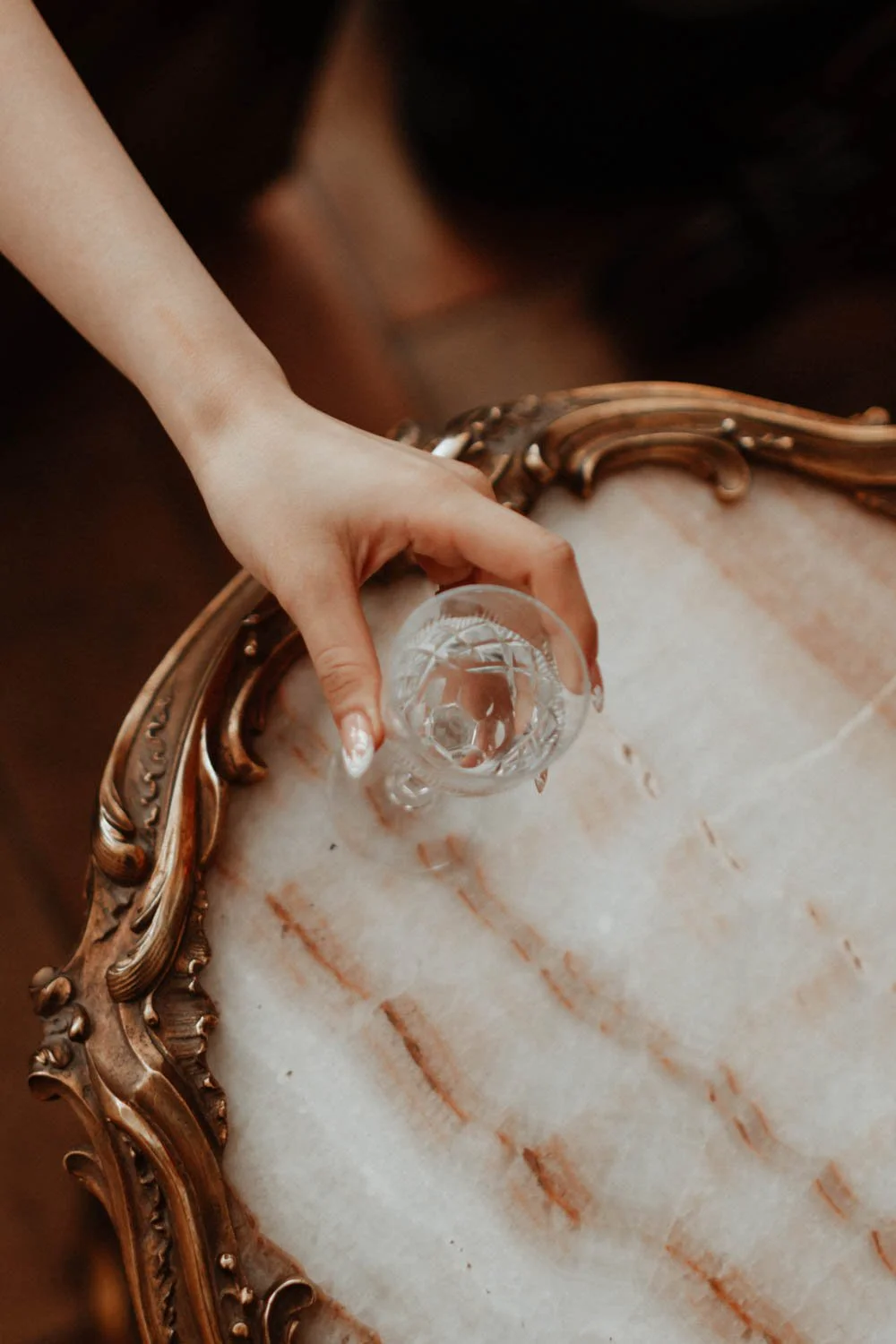 Une main verse de l'eau dans un verre en cristal sur une table en marbre ornée d'un cadre doré.