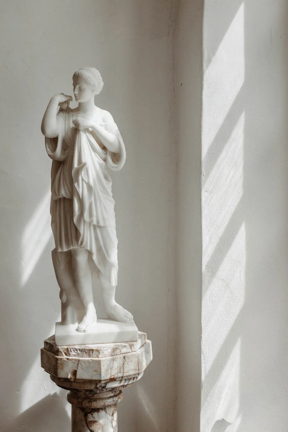 Une sculpture en marbre représentant une femme vêtue d'une robe longue, posée sur un socle en marbre, avec un rayon lumineux passant à travers une fenêtre