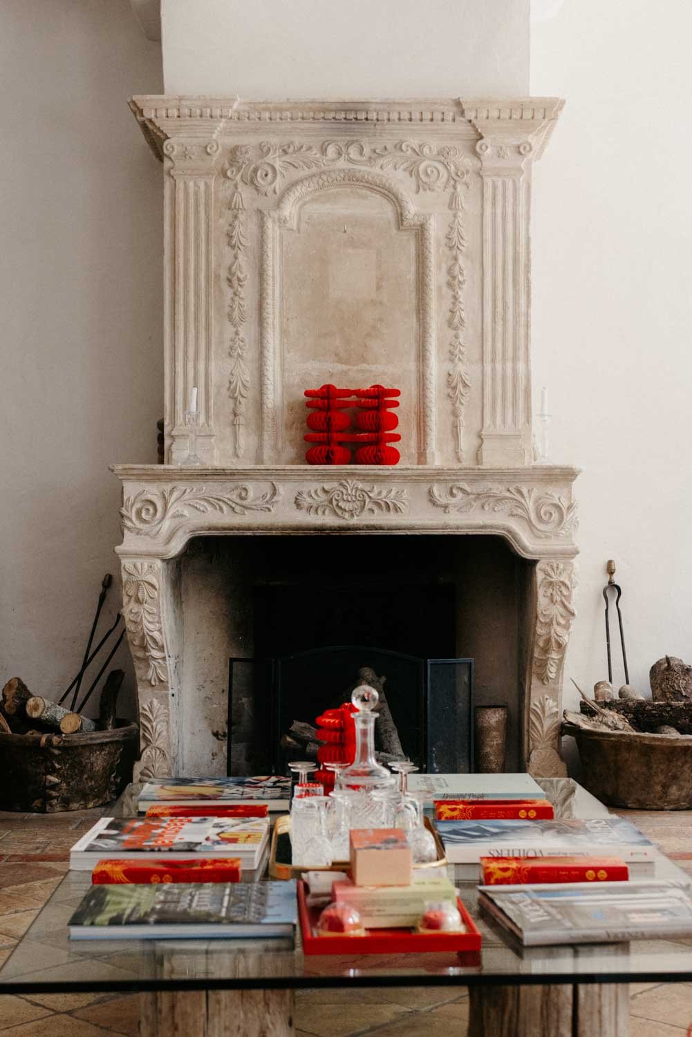 Un cheminée en pierre décorée avec des objets rouges, une table en verre avec des magazines, des livres et des objets décoratifs en verre, et deux bassines de bois remplies de bois de chauffage de chaque côté.