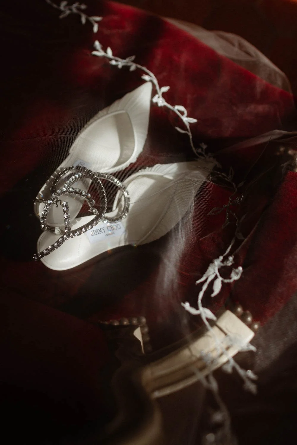 Chaussures blanches avec semelles en cuir, bijoux en forme de nœuds en cristaux, et un tag 'Jimmy Choo', disposés sur un tissu rouge foncé, avec une branche décorative en fil blanc.