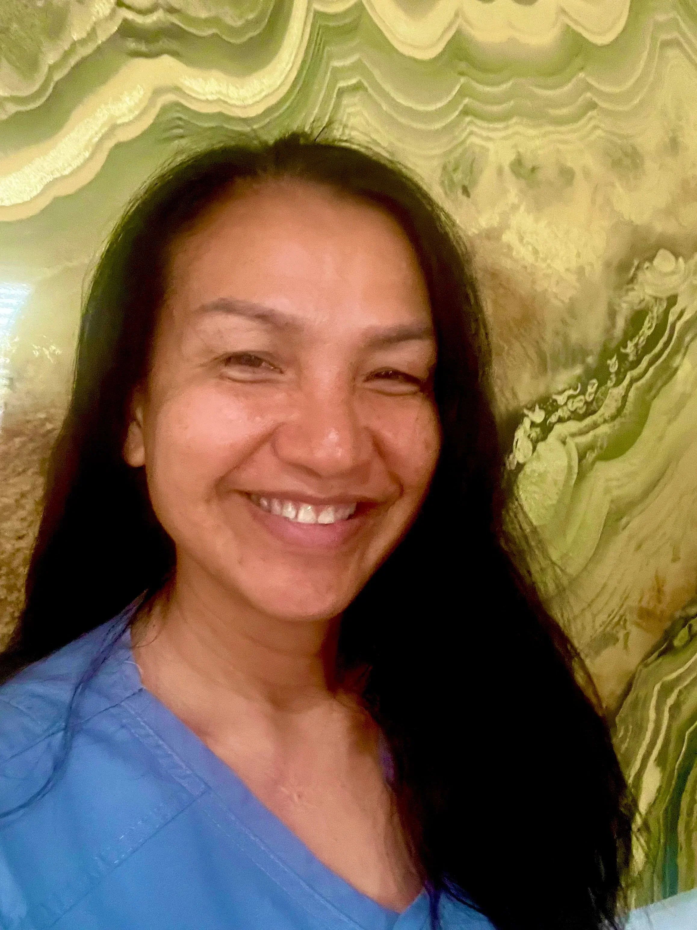 Close-up of a woman with light brown, wavy hair and a slight smile, indoors near a window with blinds.