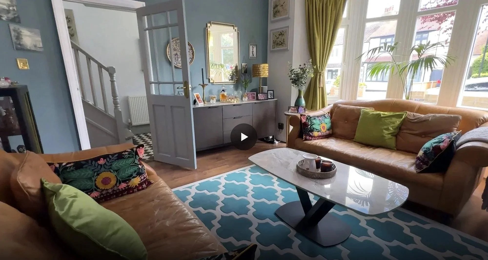 Living room with two tan leather sofas, colorful cushions, a white marble-top coffee table, a blue and white patterned rug, large windows with green curtains, potted plants, and a blue wall with frames and a mirror.