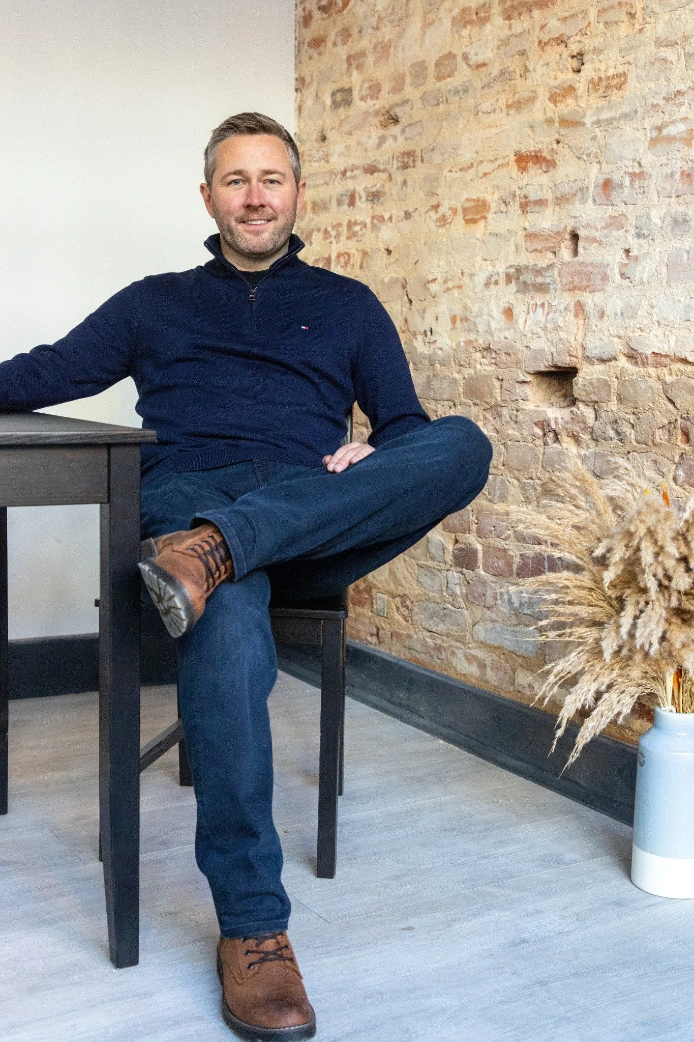 A man sitting on a chair in front of an exposed brick wall, wearing a navy sweater, jeans, and brown shoes.