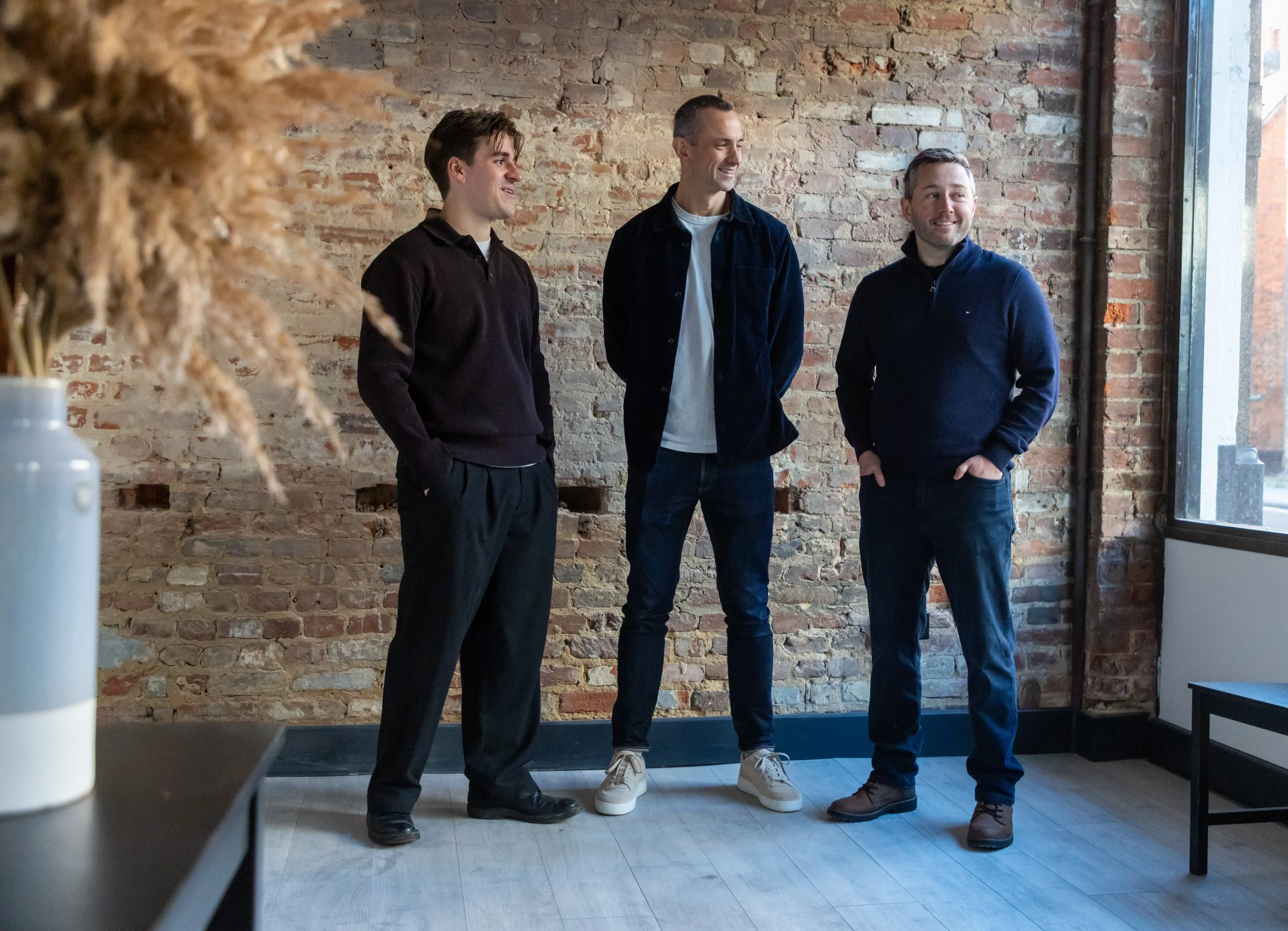 Three men standing in a row, smiling and chatting inside a room with a brick wall background and a large window on the right side.