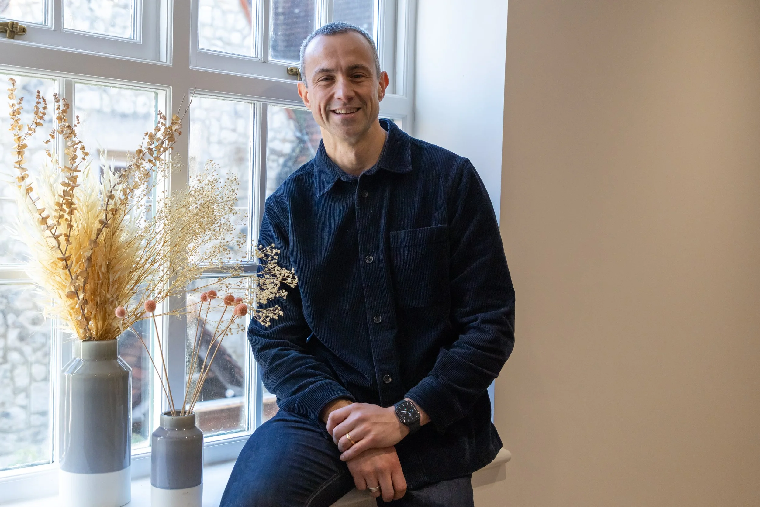 A man sitting on a windowsill, smiling, wearing a dark blue shirt and a watch, with two vases of dried flowers on his left.