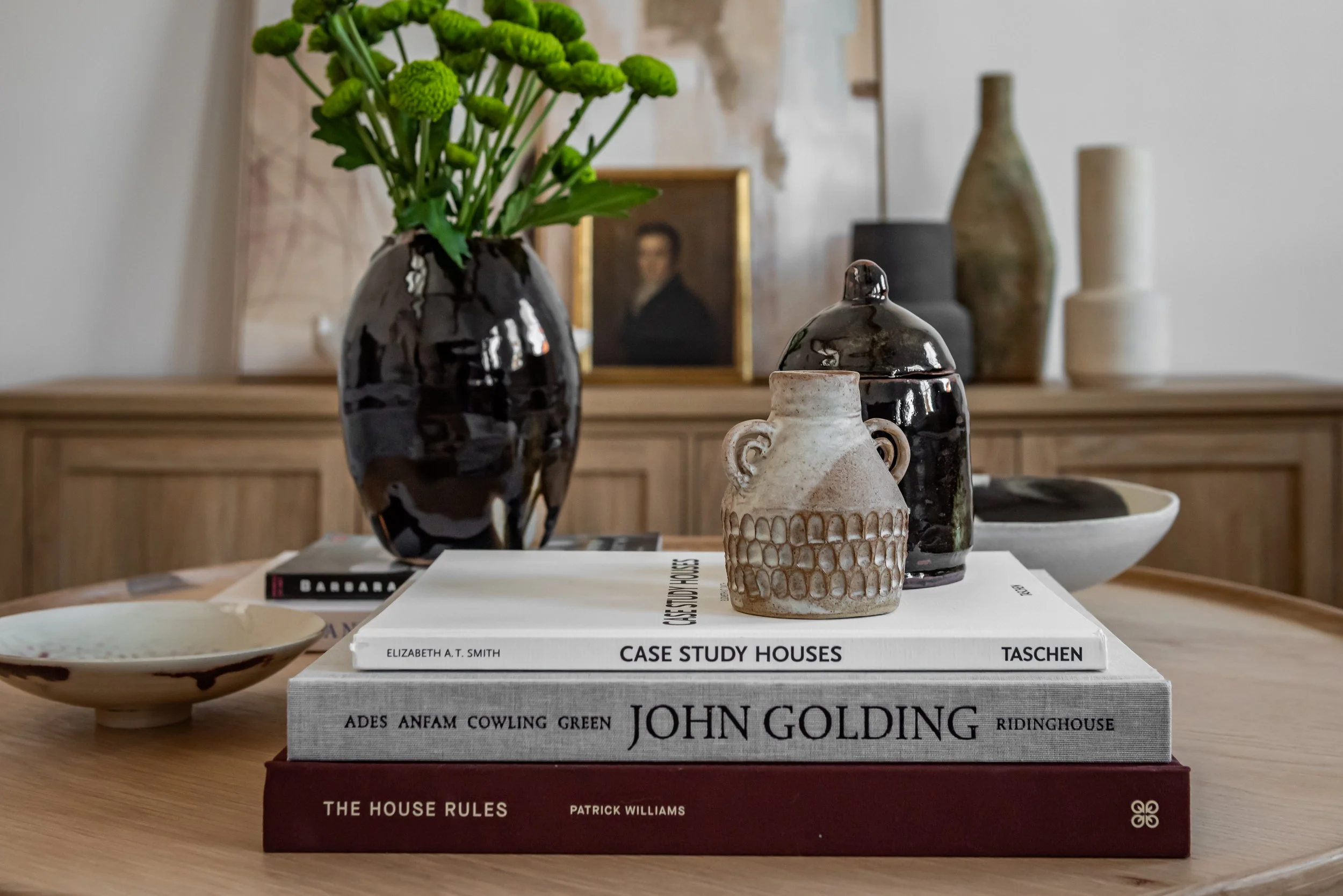 A decorative arrangement on a wooden table including a black ceramic vase with green flowers, three ceramic jars, a small dish, and two stacked books titled 'CASE STUDY HOUSES' by Elizabeth A.T. Smith and 'THE HOUSE RULES' by Patrick Williams with a large book by John Golding underneath. In the background, there are vases and a framed portrait on a wooden sideboard.
