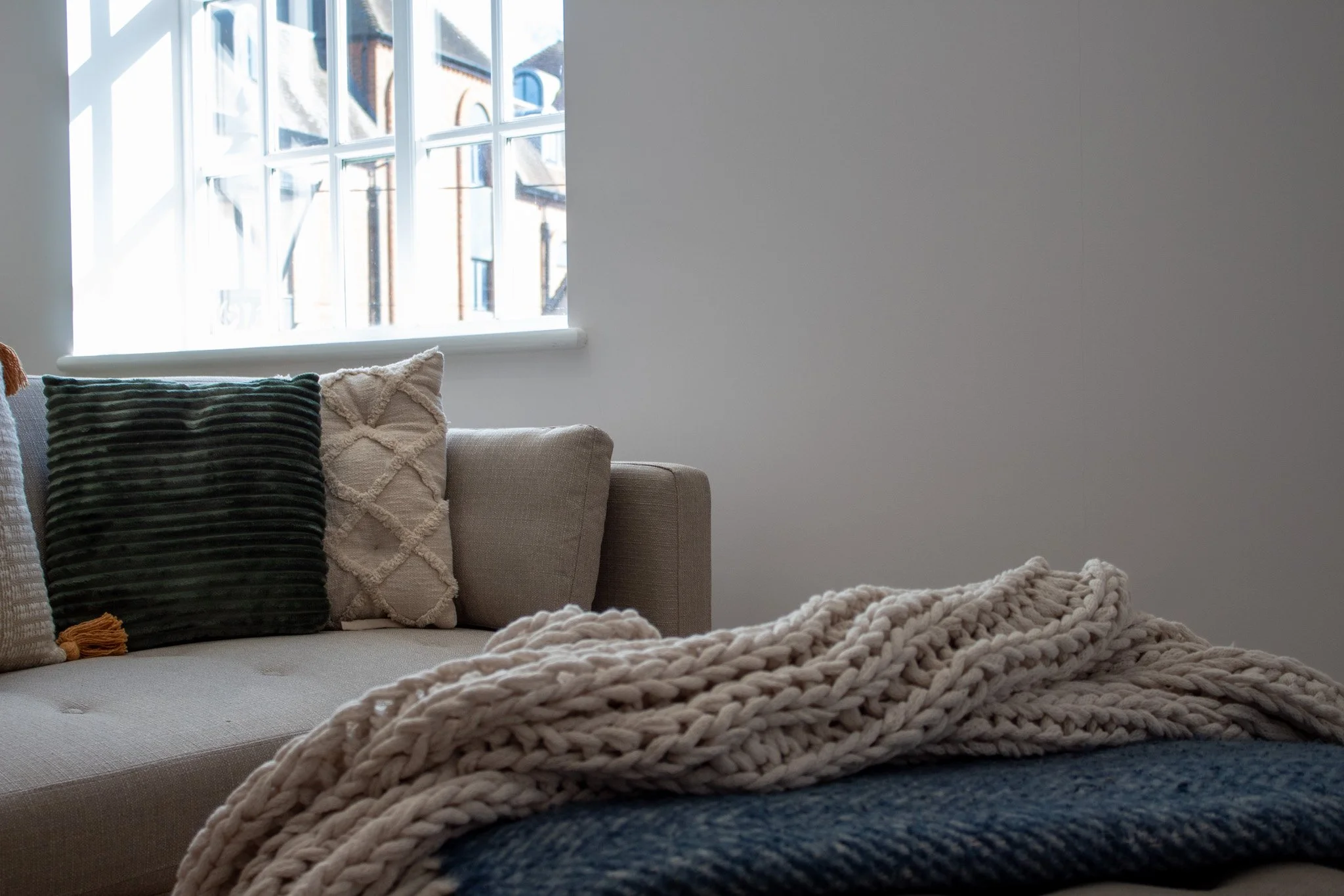 A cozy living room corner with a beige sofa, decorative pillows including a dark green velvety pillow and a cream pillow with a lattice design, a chunky knitted blanket, and a window showing a view of neighboring buildings.
