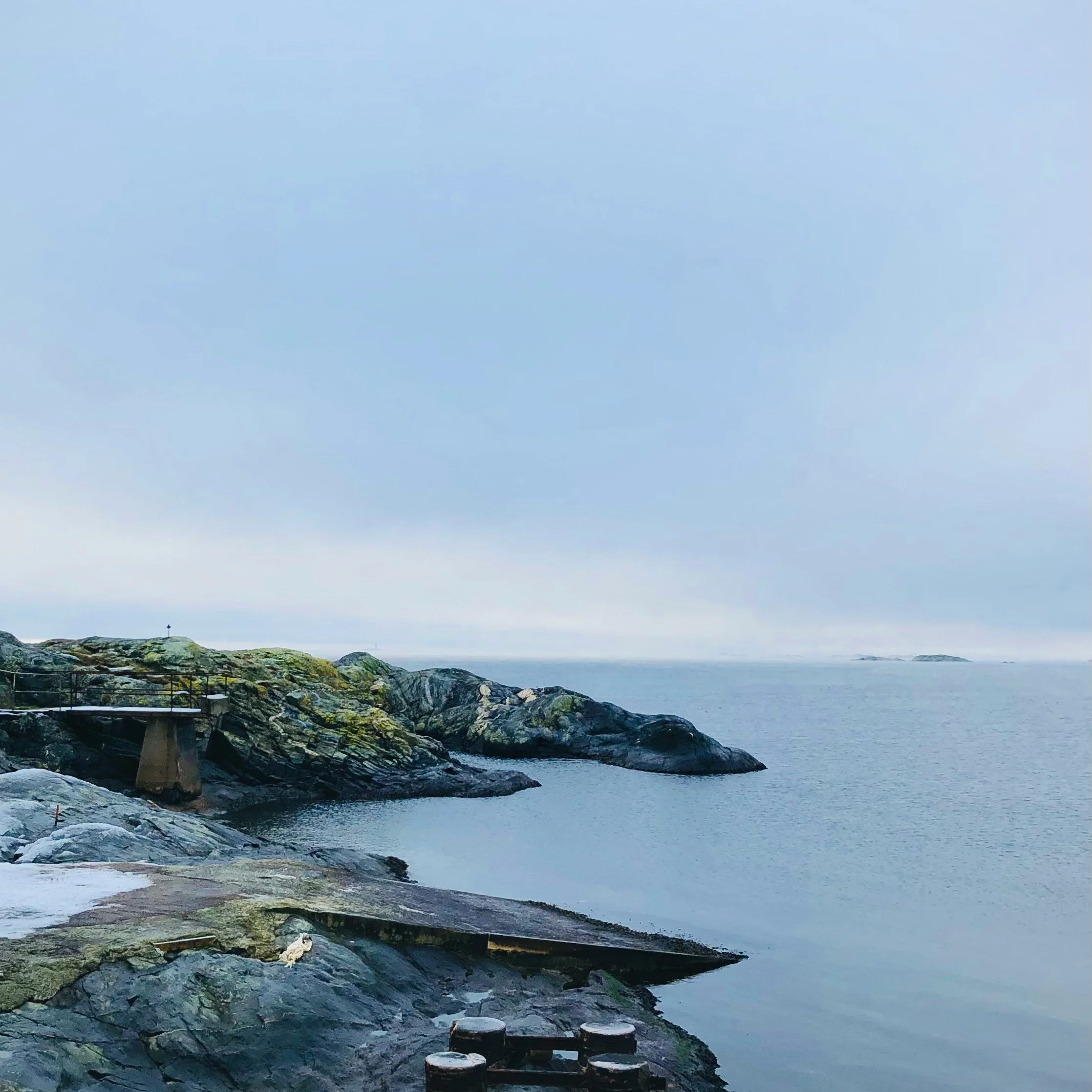 Kustlinje med vågor som slår mot klippor och en liten brygga, under en molnig himmel.