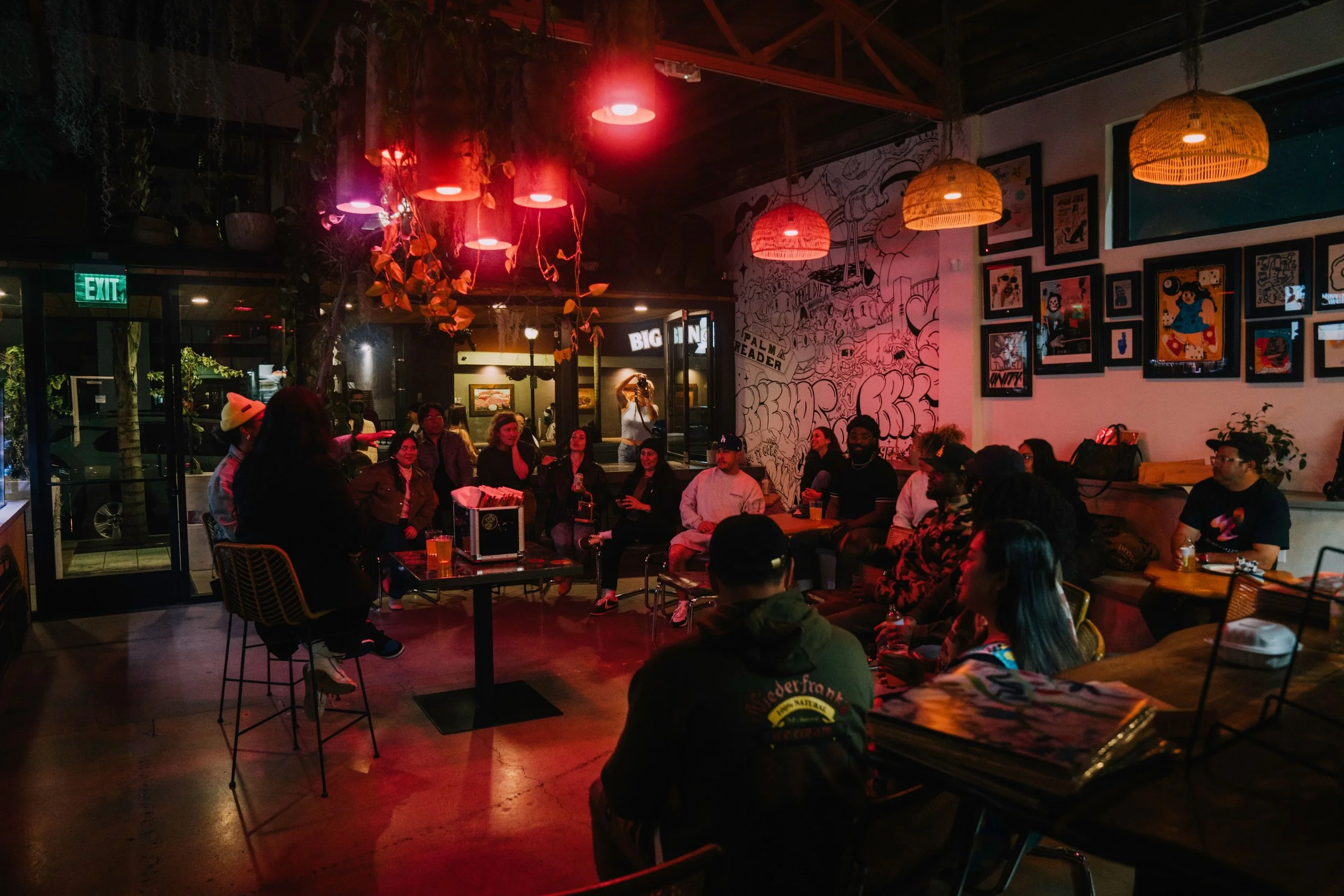 People gathered in a dimly lit indoor venue with hanging lights and wall art, sitting around tables, some holding drinks, engaged in a group conversation or listening to a speaker.