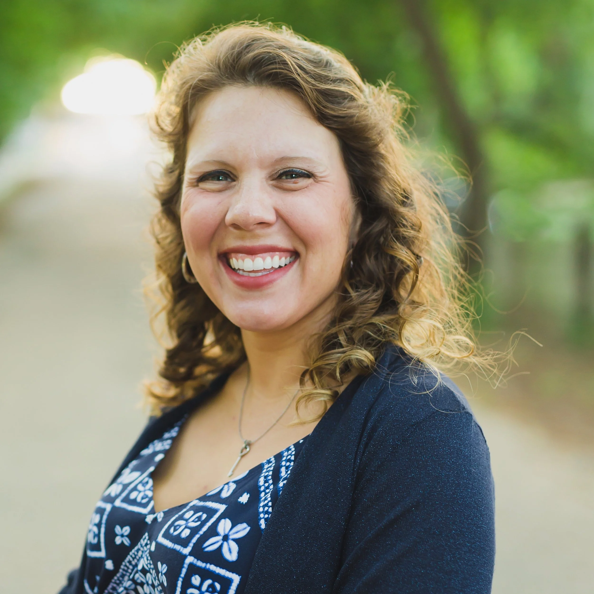A smiling woman with curly brown hair wearing a dark blazer and a patterned top outdoors with green trees in the background.
