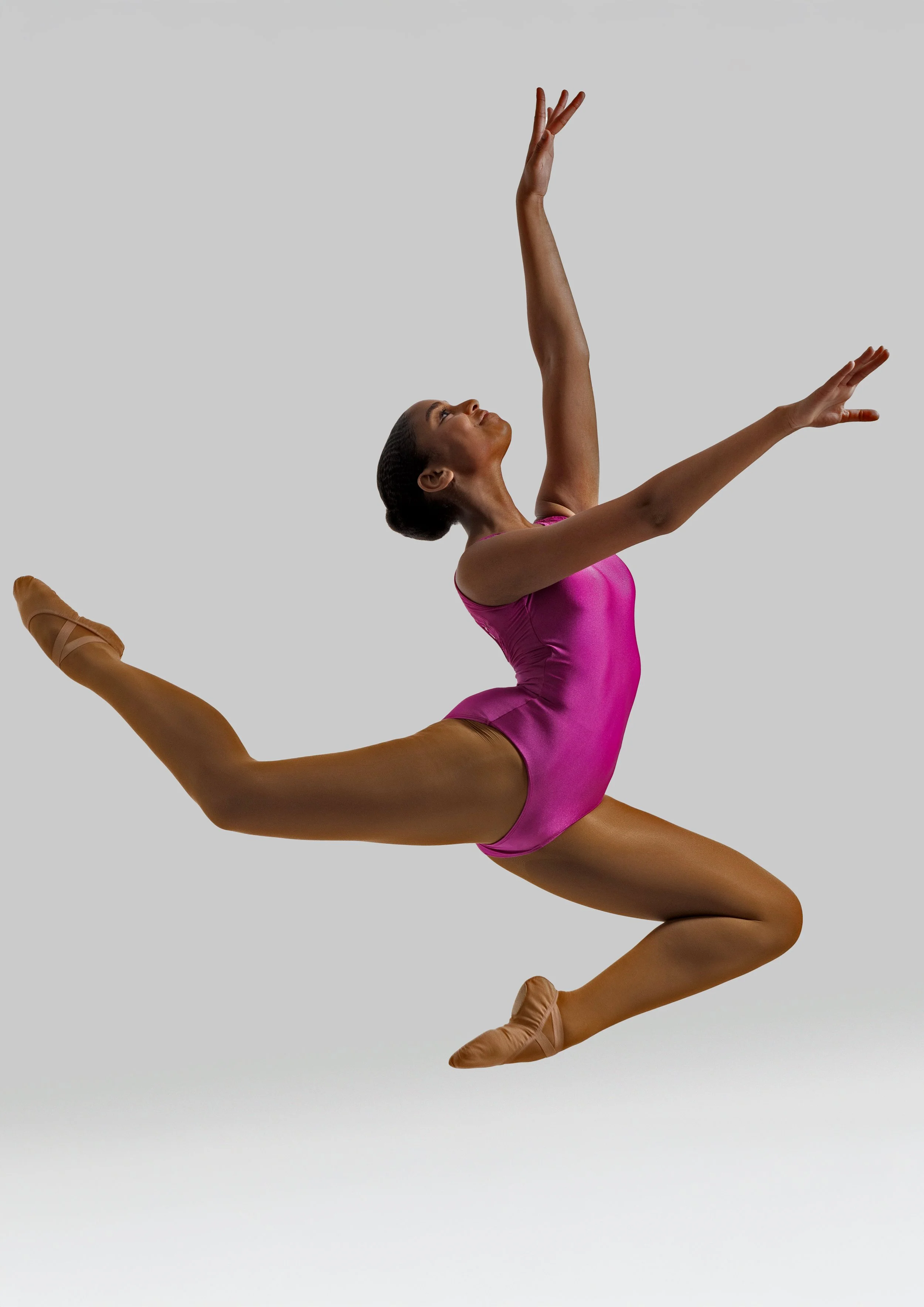 A female gymnast wearing a pink leotard performing a leap in mid-air against a plain gray background.