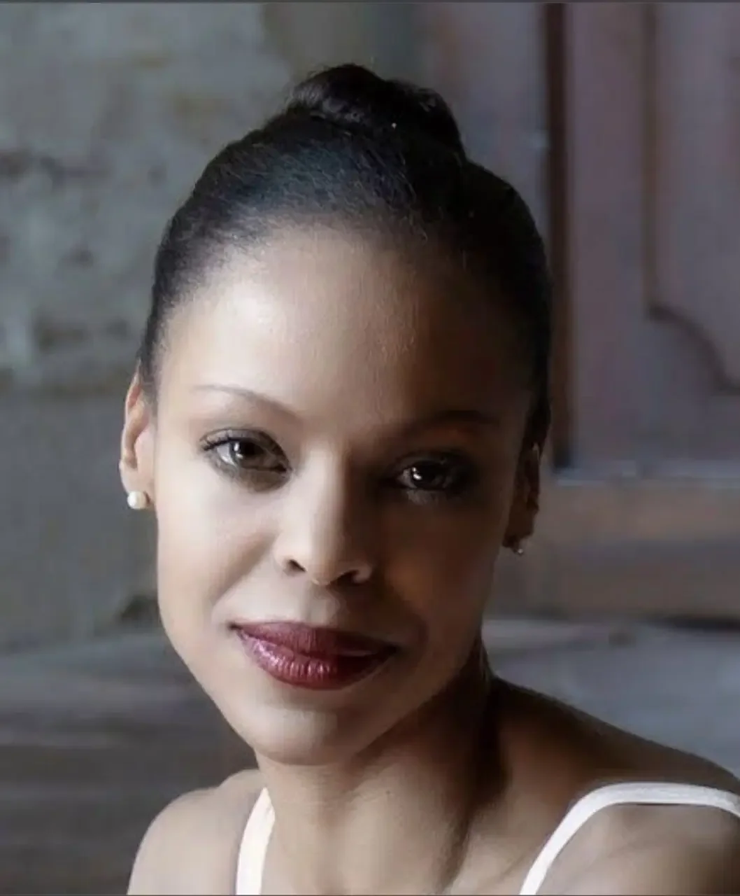 Close-up of a woman with her hair in a bun, wearing pearl earrings and red lipstick, looking at the camera with a slight smile.