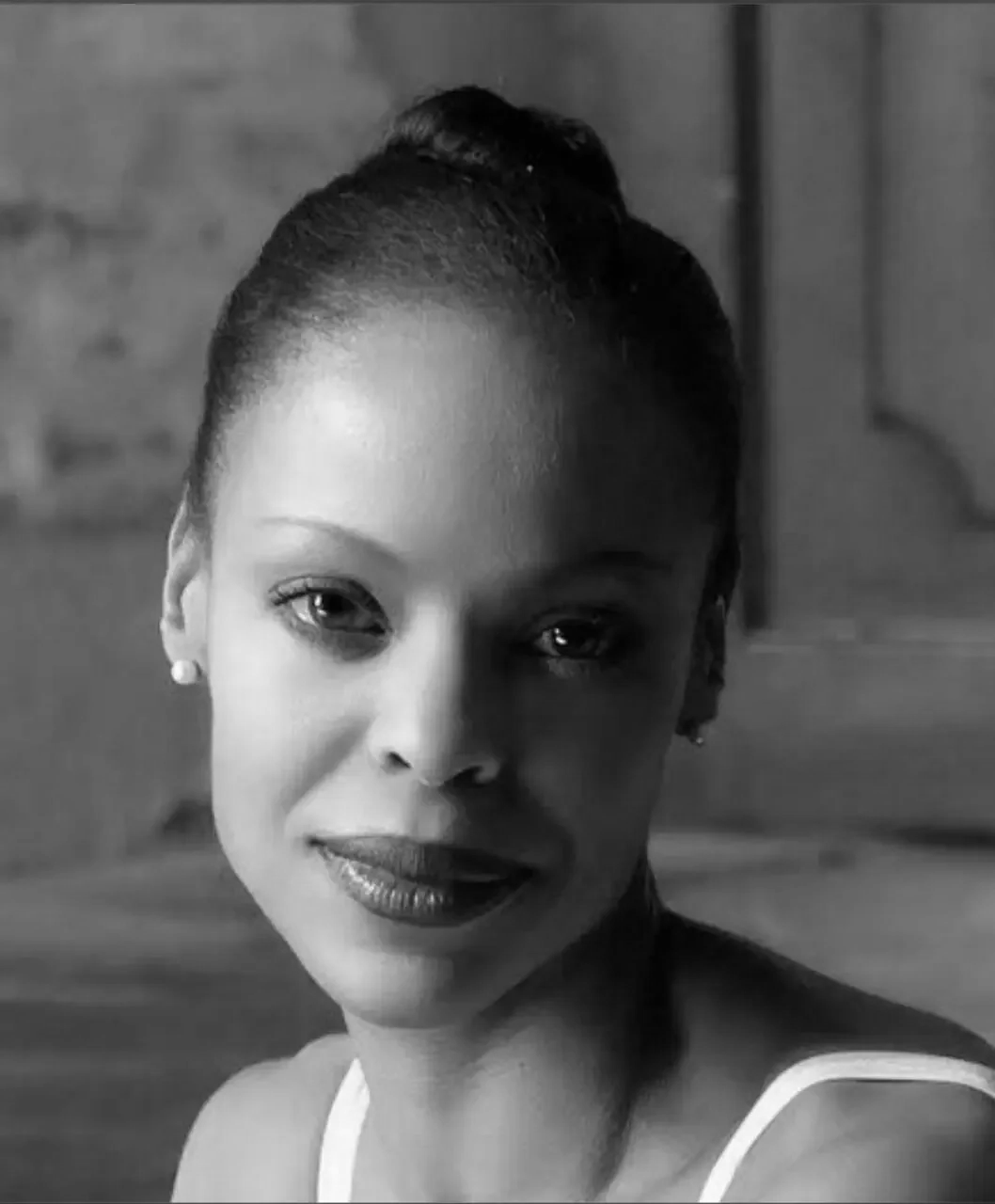Black and white portrait of a woman with her hair styled in a bun, wearing pearl earrings and a sleeveless top, looking directly at the camera.
