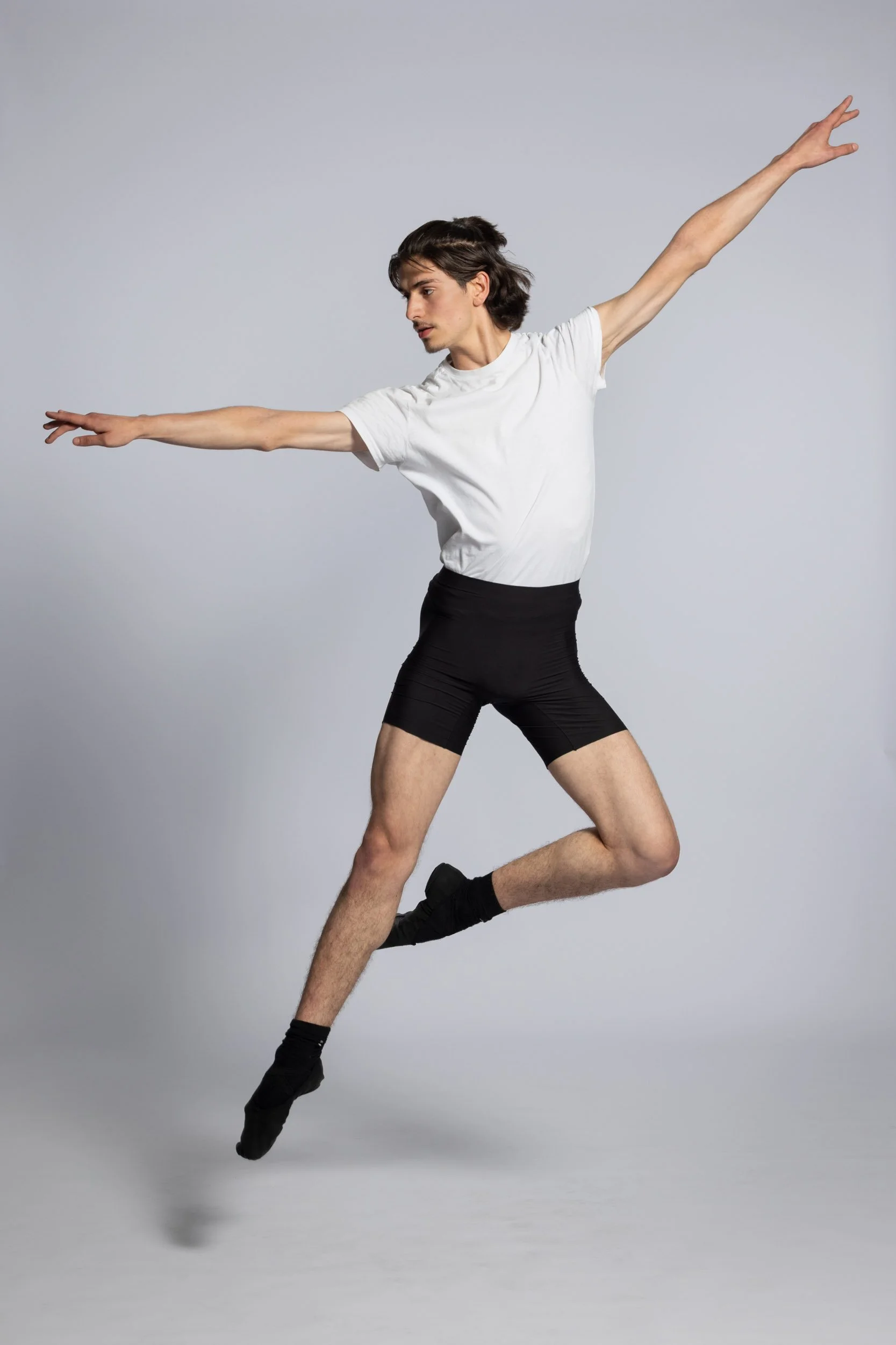 A male dancer in a T-shirt and shorts is captured mid-air performing a dance move against a plain gray background.