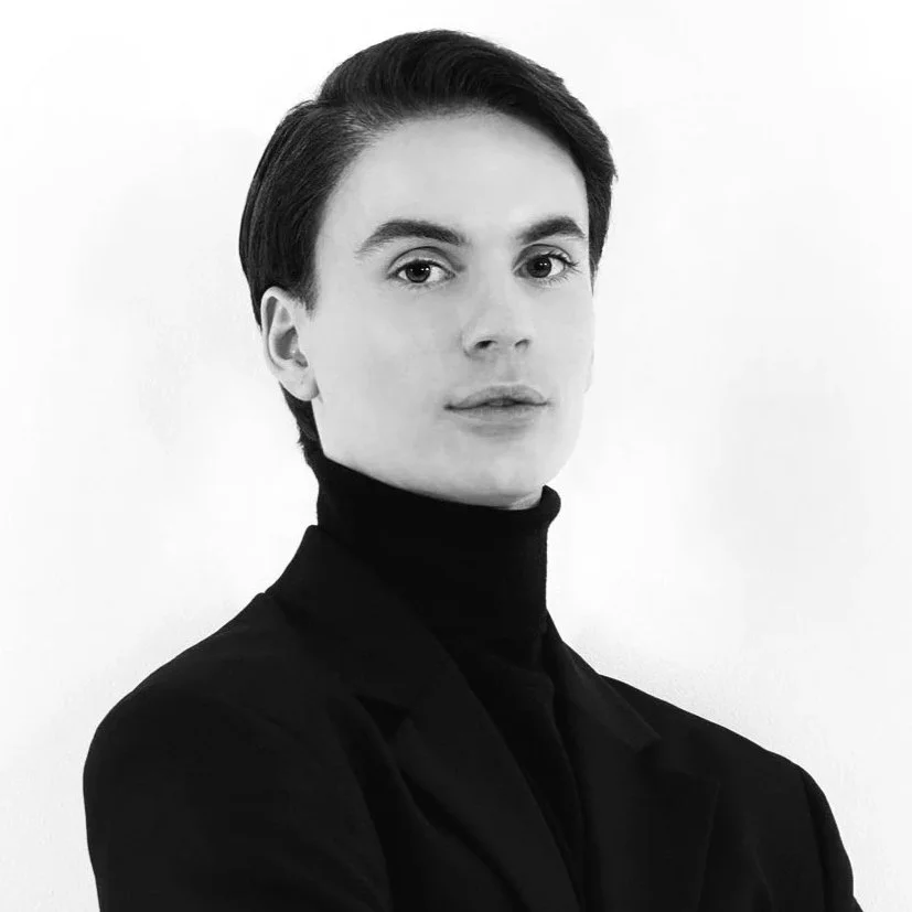 Black and white portrait of a young woman with short dark hair, wearing a black turtleneck and blazer, posing against a plain white background.