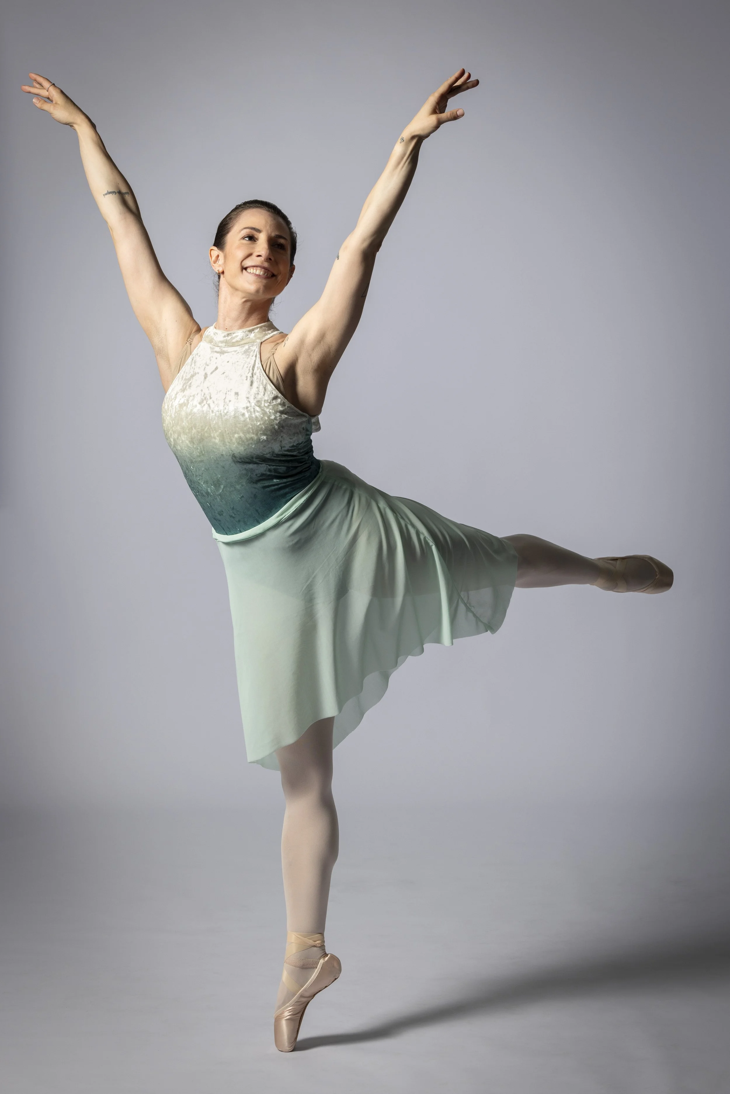 A woman in a dance pose, standing on one foot in ballet slippers, with arms raised and a smile, wearing a gradient sleeveless top and a light green skirt, against a plain gray background.