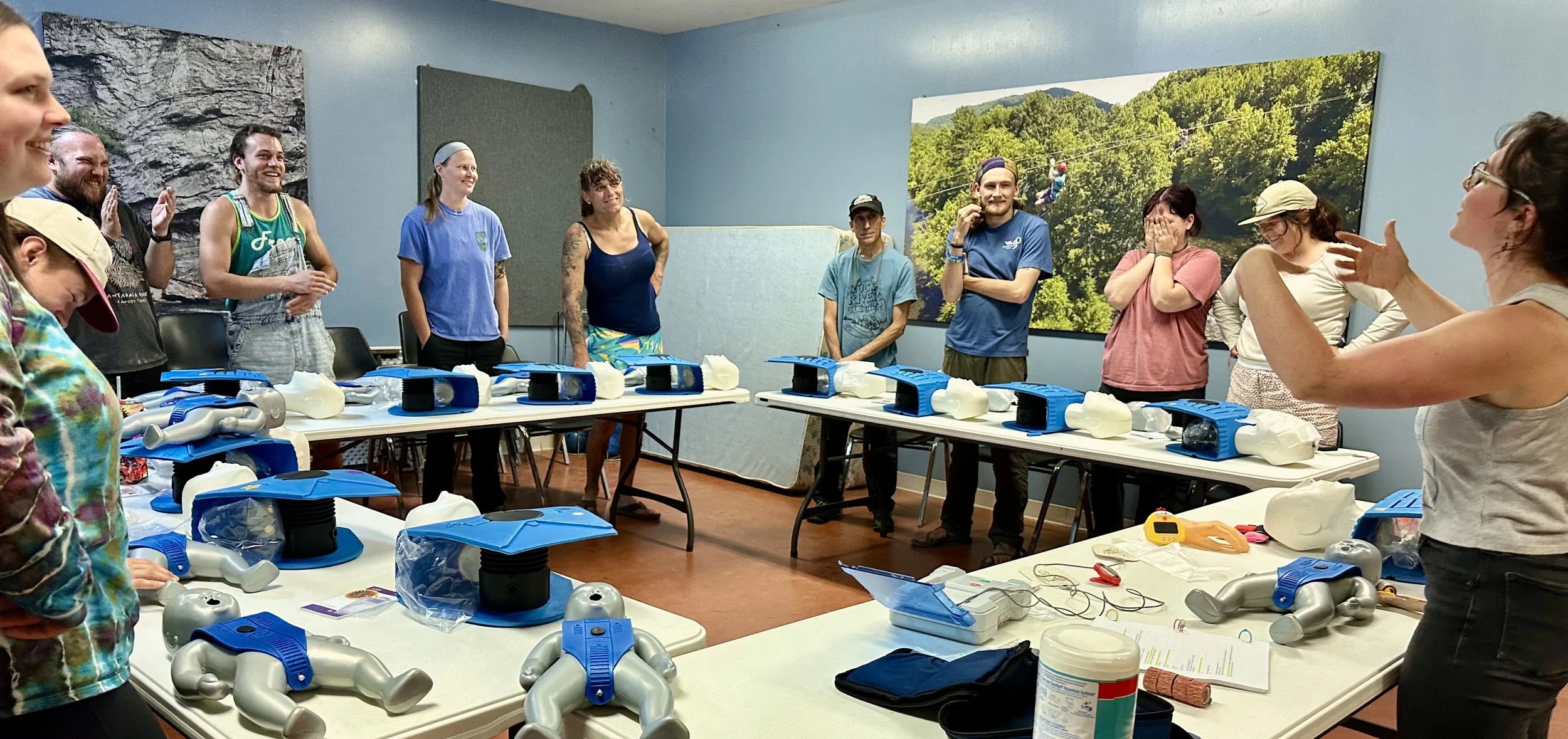 Group of people standing around tables with CPR mannequins and medical equipment in a classroom or training room.