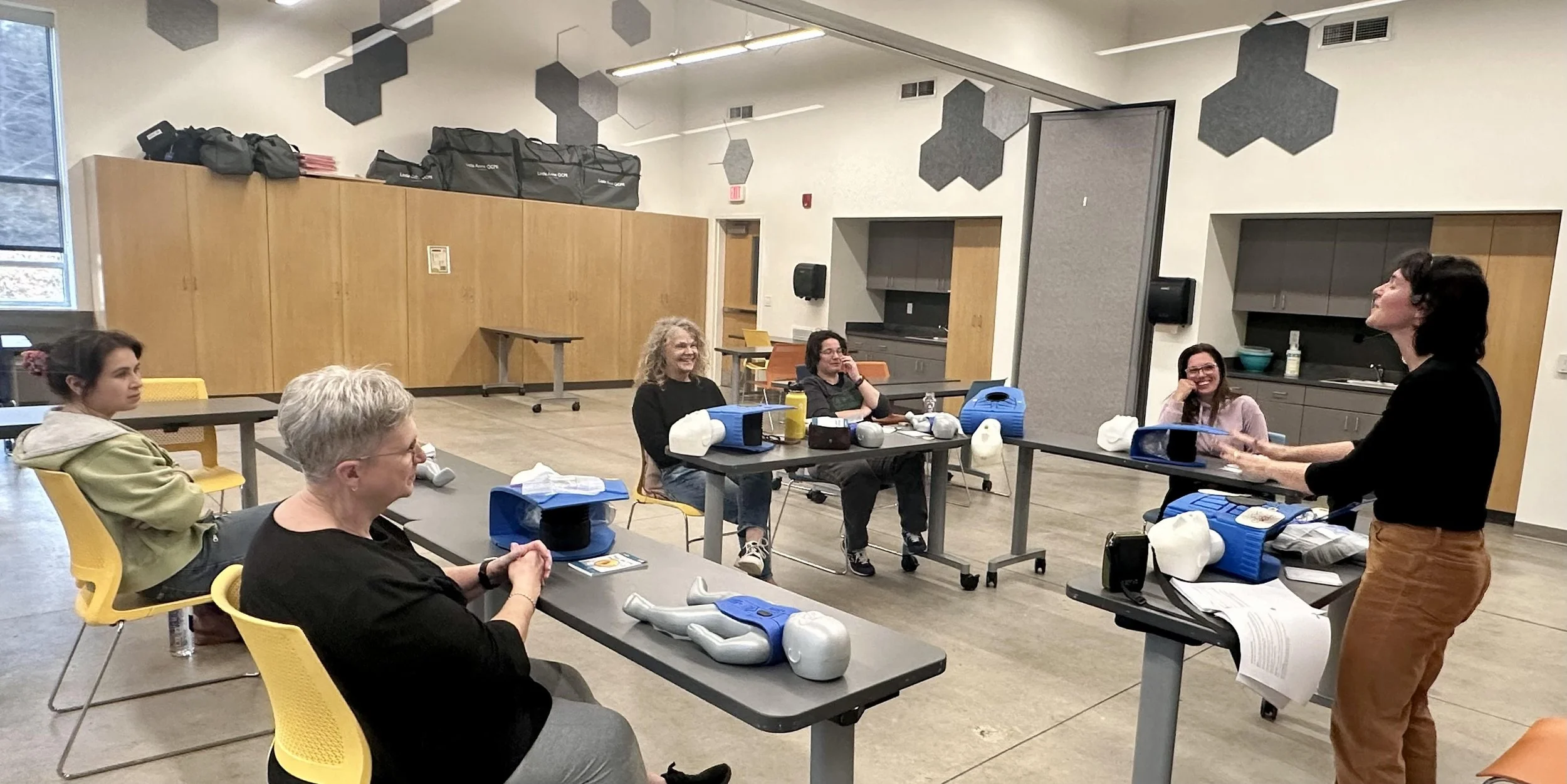 A group of people participating in a CPR training course, sitting at tables with manikins and training equipment, listening to an instructor standing at the front of the room.