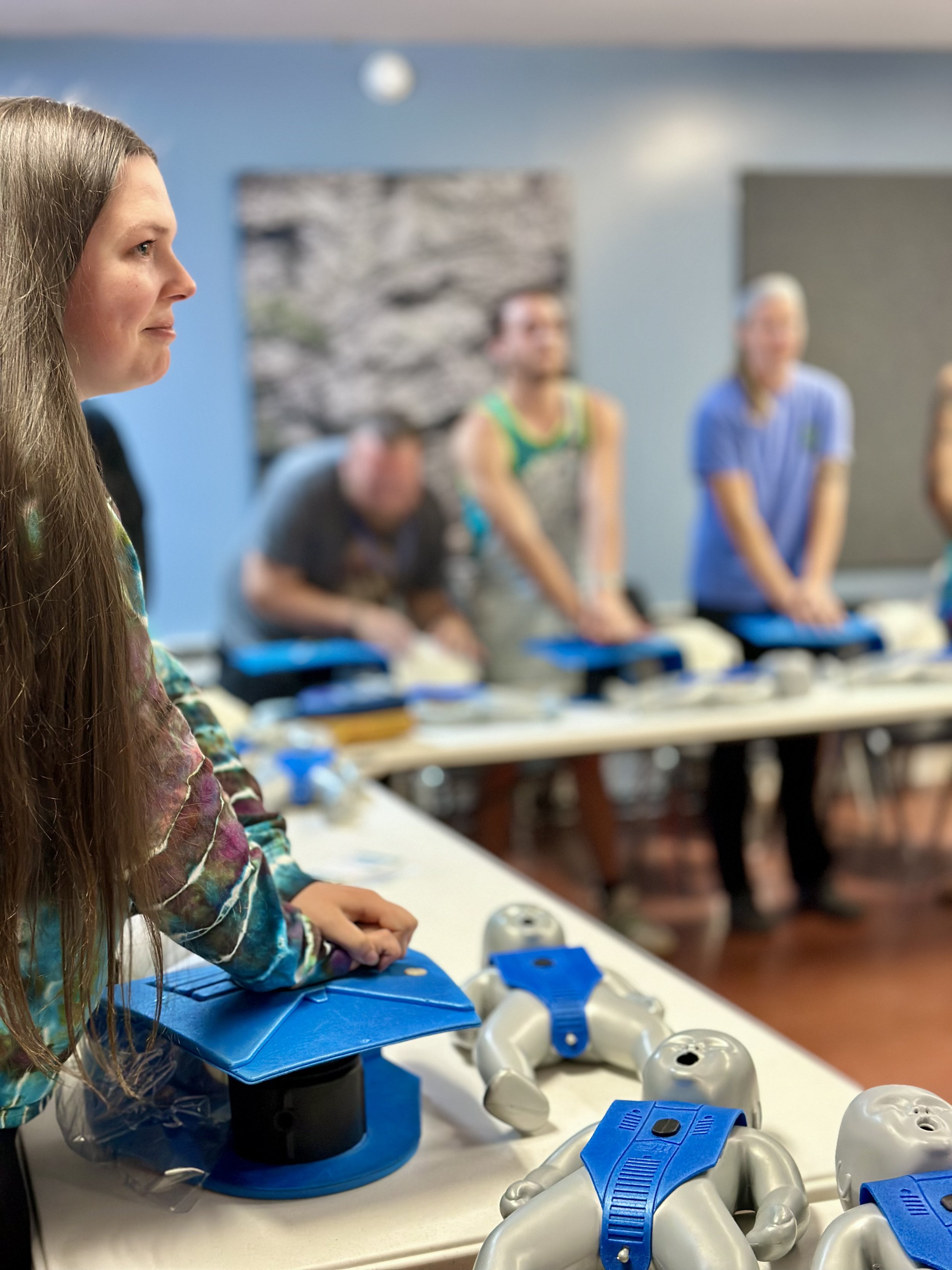 A woman with long blonde hair sits at a table with medical training models and equipment, with several other people in the background in a classroom or training setting.