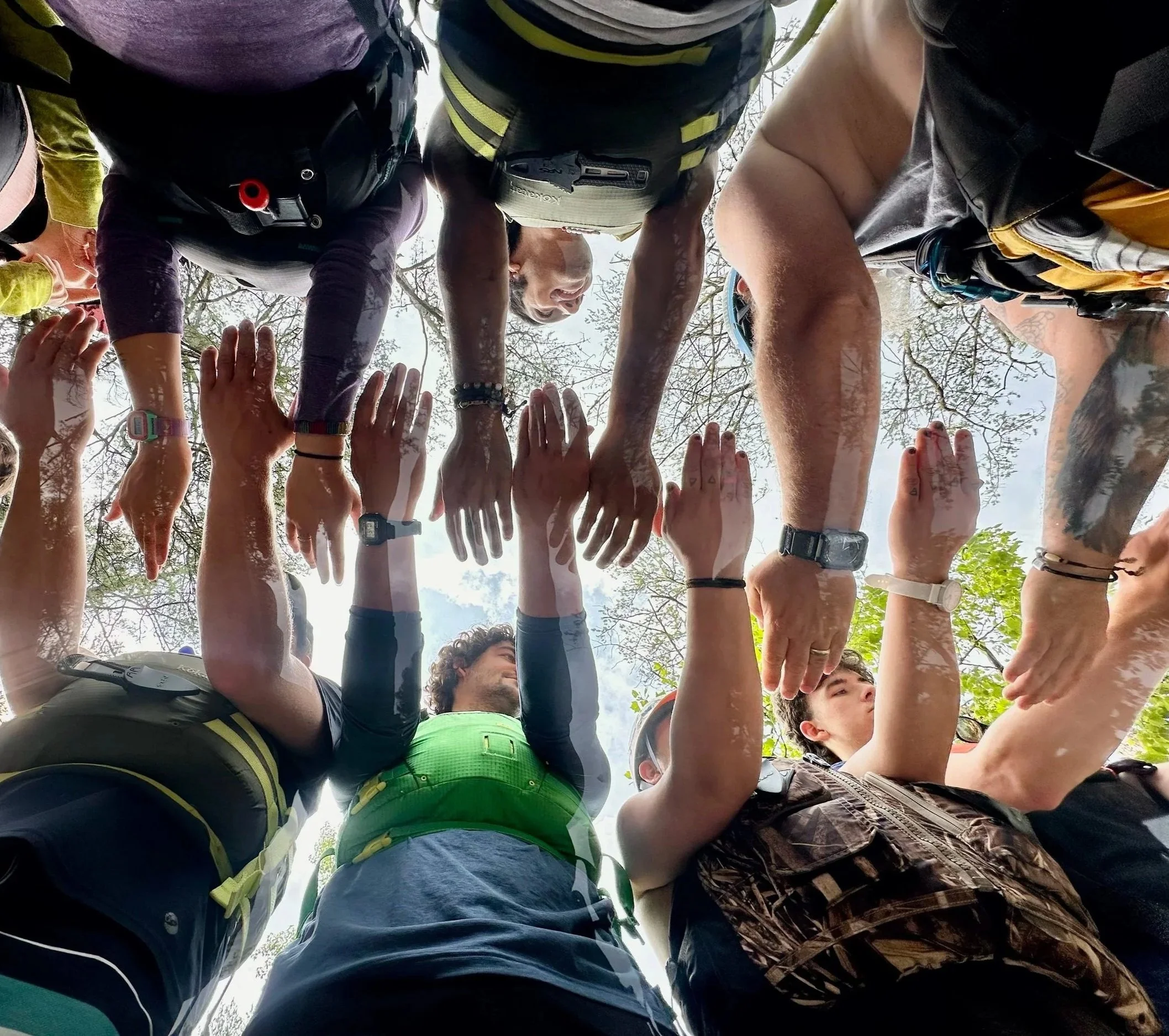 People forming a human pyramid outdoors on a bright, cloudy day, reaching downward with their hands to support each other.