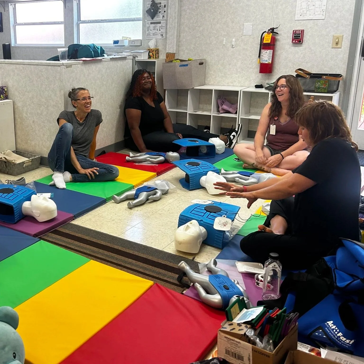 Group of five women sitting on colorful mats on the floor, playing with medical training mannequins in a classroom or training room setting. They are smiling and appear to be engaged in a hands-on activity.