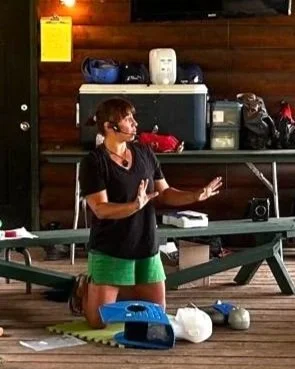 Woman wearing a headset giving a presentation in front of a volleyball net, with a volleyball and other sports equipment on the floor.