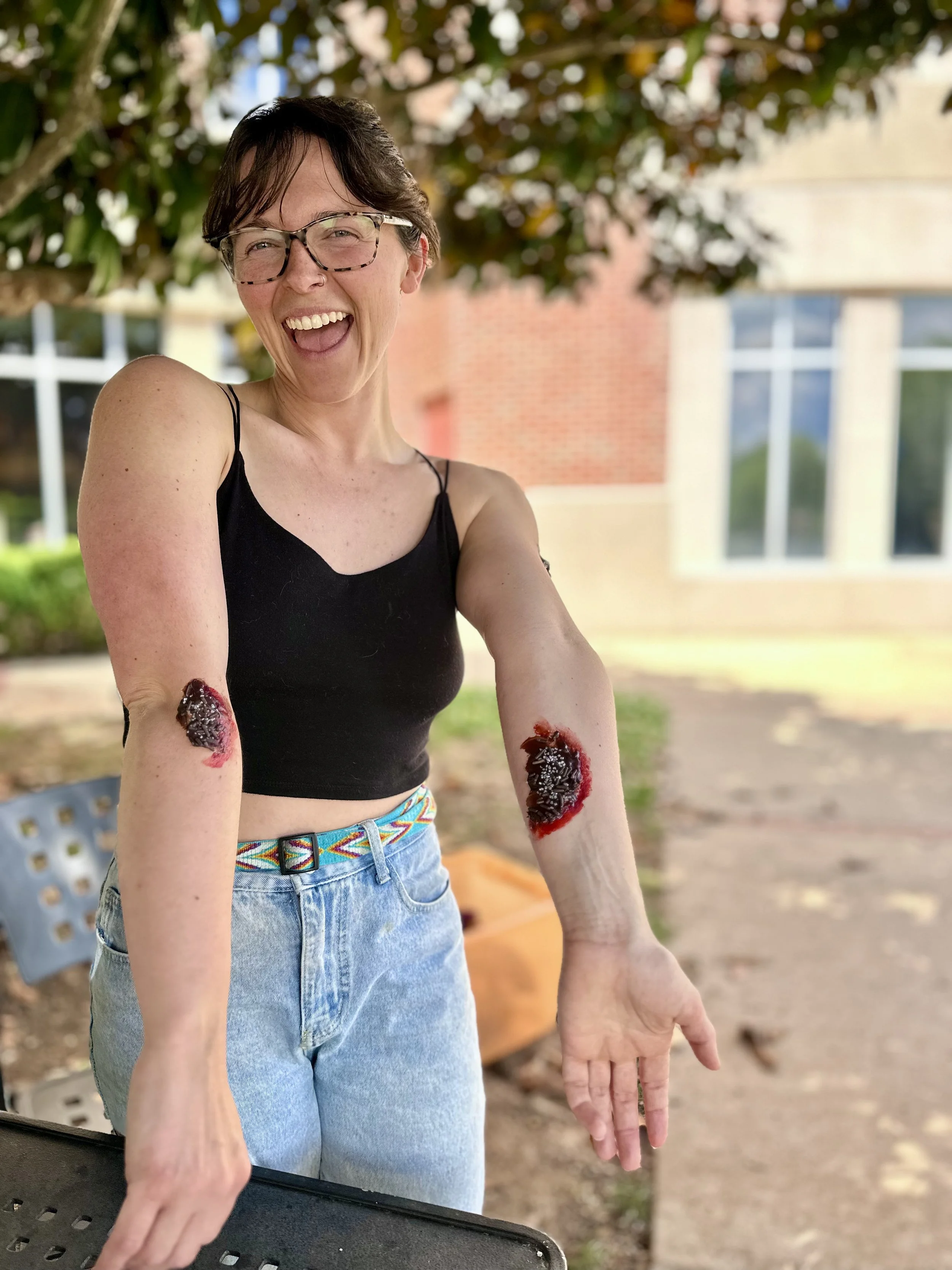 A woman with short dark hair, glasses, and a black tank top laughing outdoors, showing her arm with snake bites covered in fake blood and glittery decorations on her forearms, in front of a building and trees.