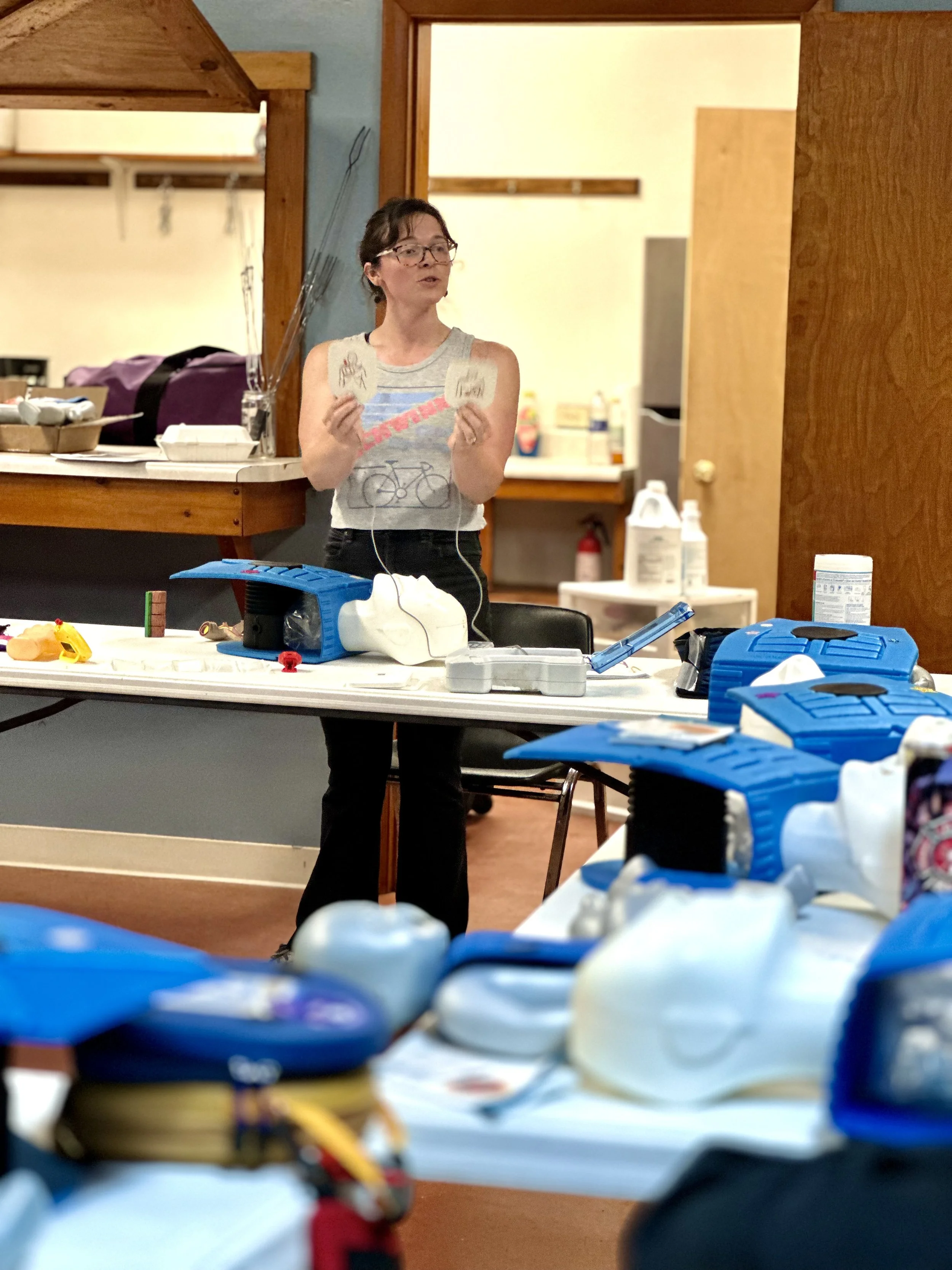 Mita Coker is wearing glasses and a sleeveless shirt is holding some cards and speaking, standing behind a table with various medical or surveillance devices and supplies in a room with wooden accents and a kitchen in the background.