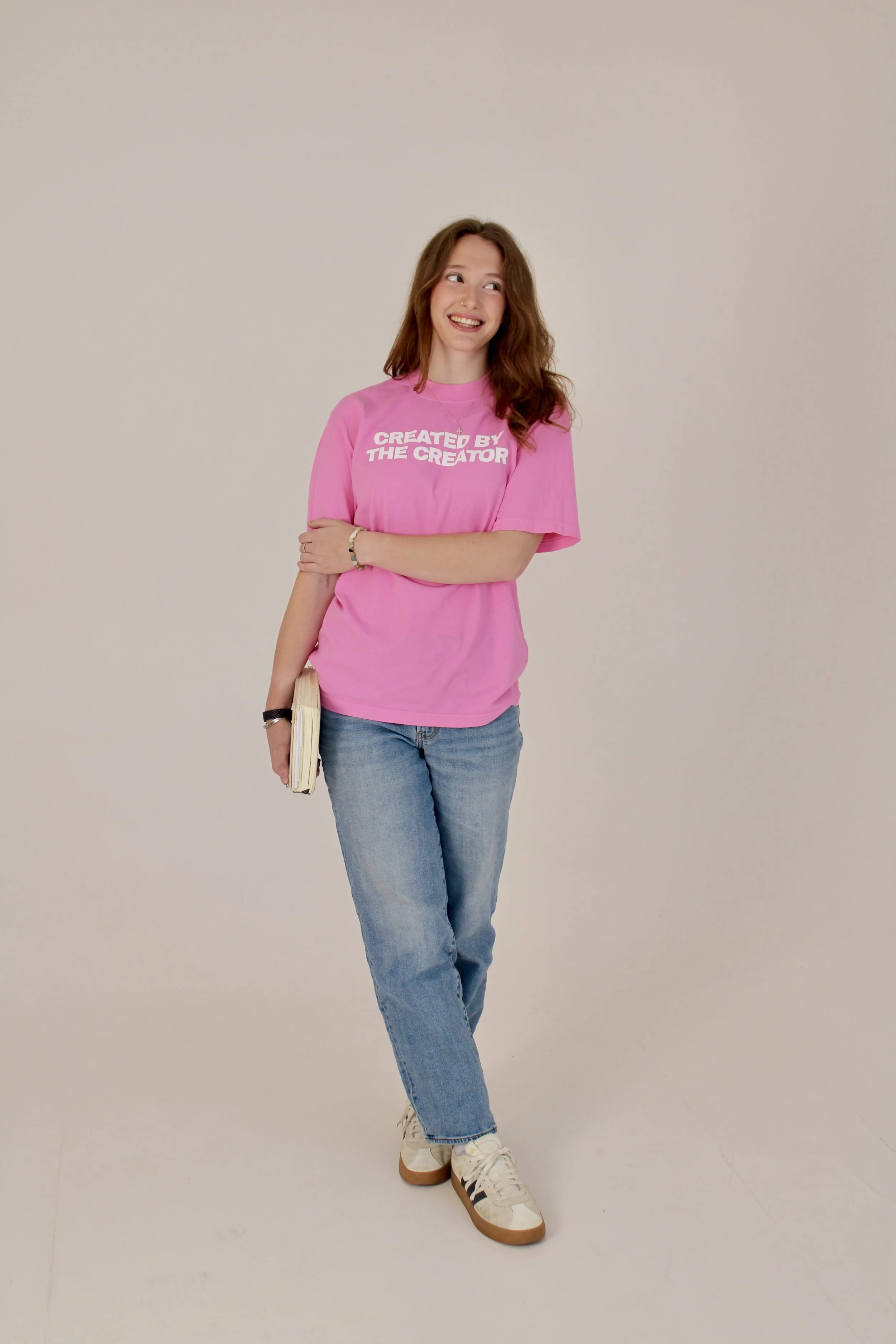 A young woman with red hair wearing a pink t-shirt with white text that says 'CREATED BY THE CREATOR'. She is standing against a plain off-white background, smiling and looking to her right. She has a bracelet on her right wrist, is holding a book or notebook in her left hand, and is wearing blue jeans and white sneakers.