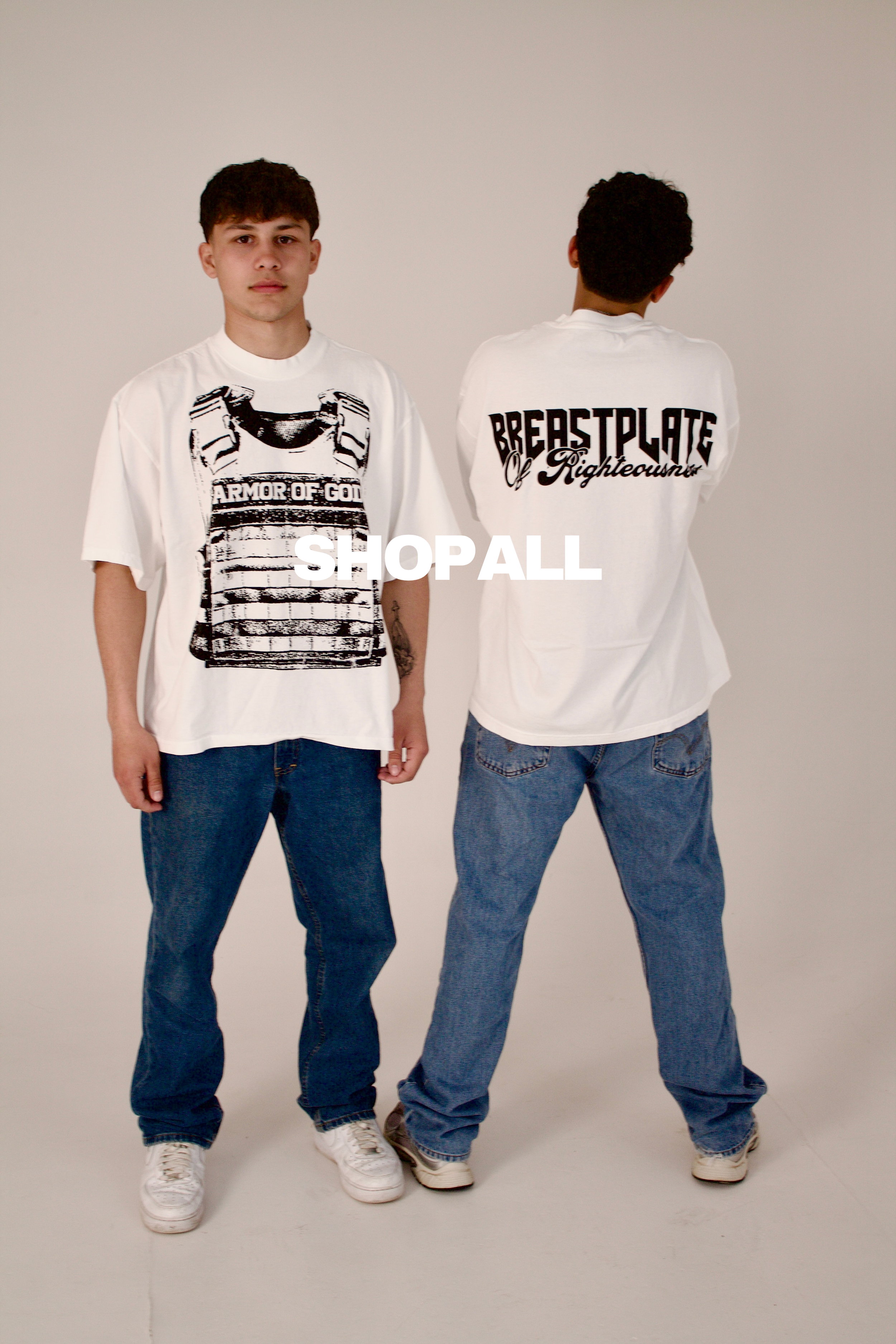 Two young men wearing white graphic T-shirts and blue jeans standing against a plain wall, one facing forward and the other with his back to the camera.