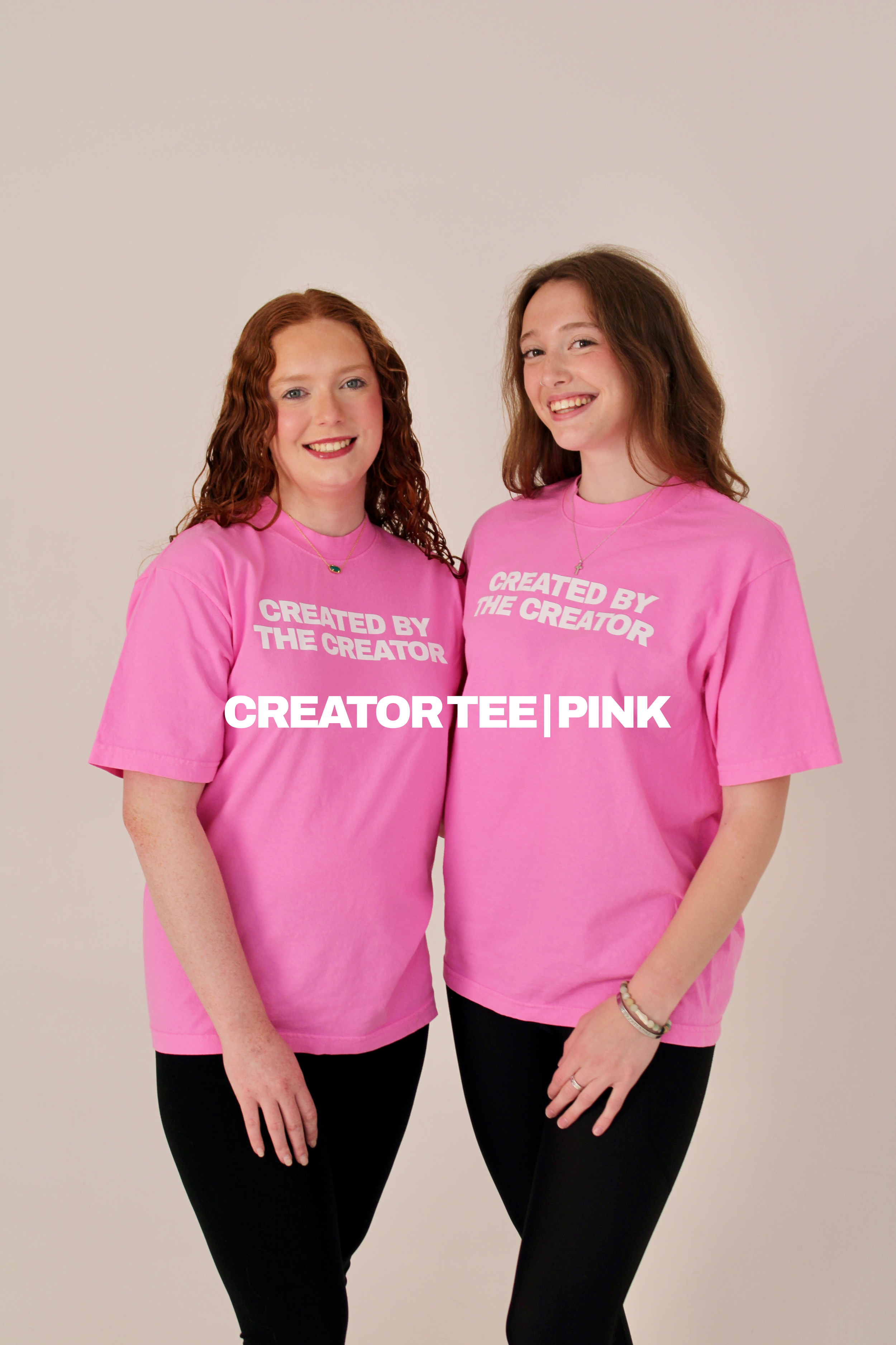 Two women standing side by side wearing pink T-shirts with the words "Created by the Creator" printed on them, smiling against a plain background.