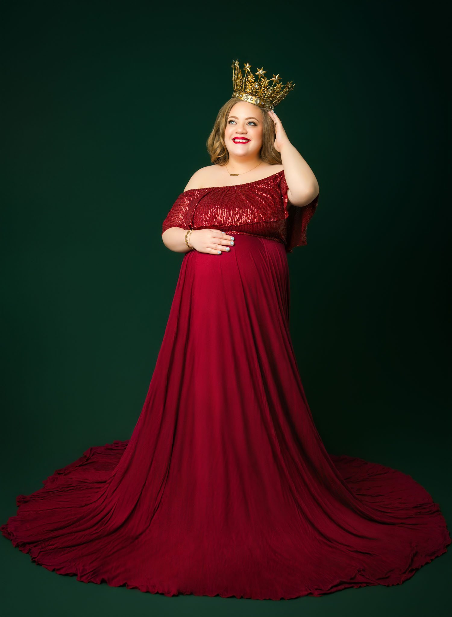 Pregnant woman in a dramatic red gown posing against a deep green studio backdrop.
