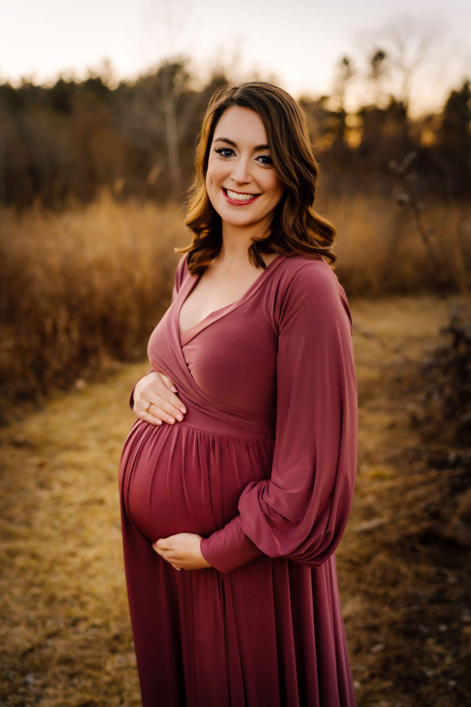 Expecting mother smiling in a mauve dress while cradling her baby bump in a field.