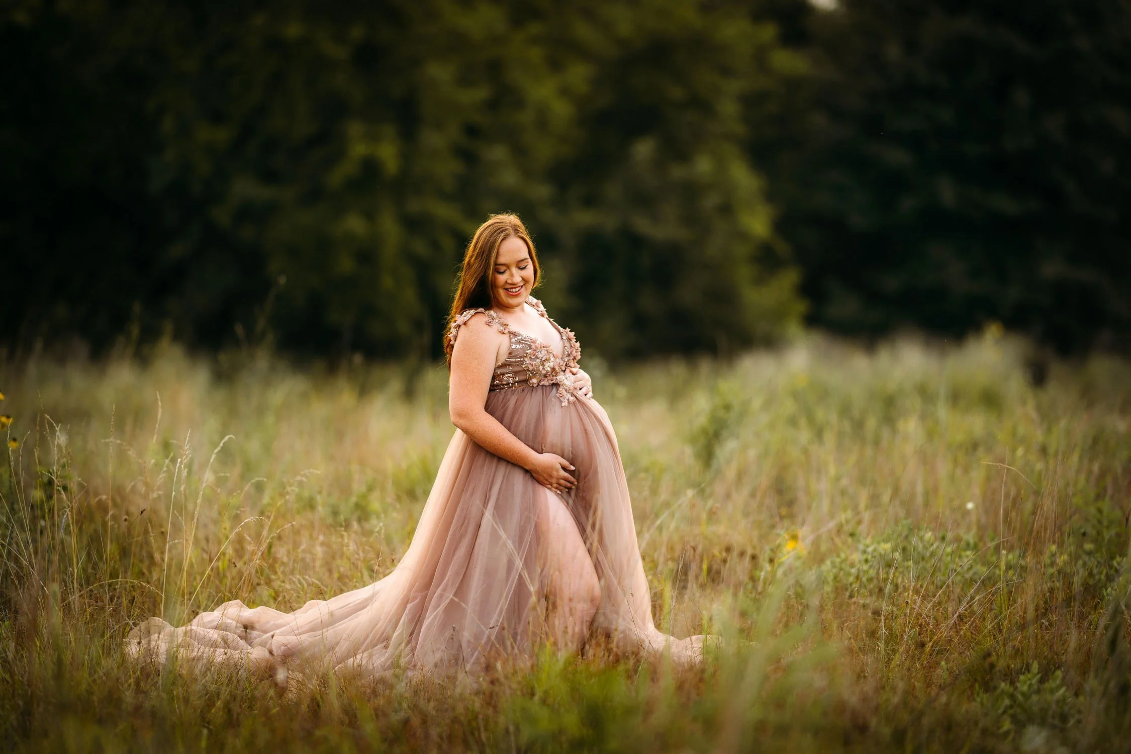 Expecting mother in a blush gown sitting in a grassy field during an outdoor maternity portrait.