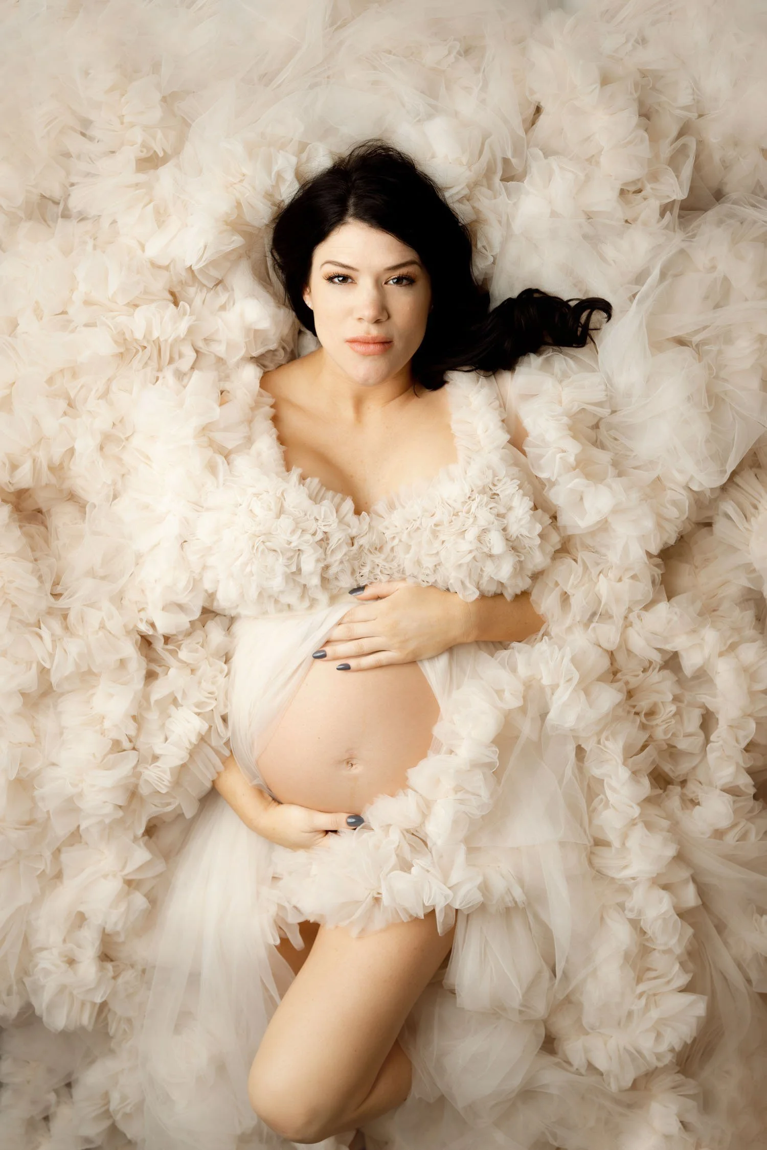 Expecting mother lying among soft cream dress in an elegant maternity portrait.