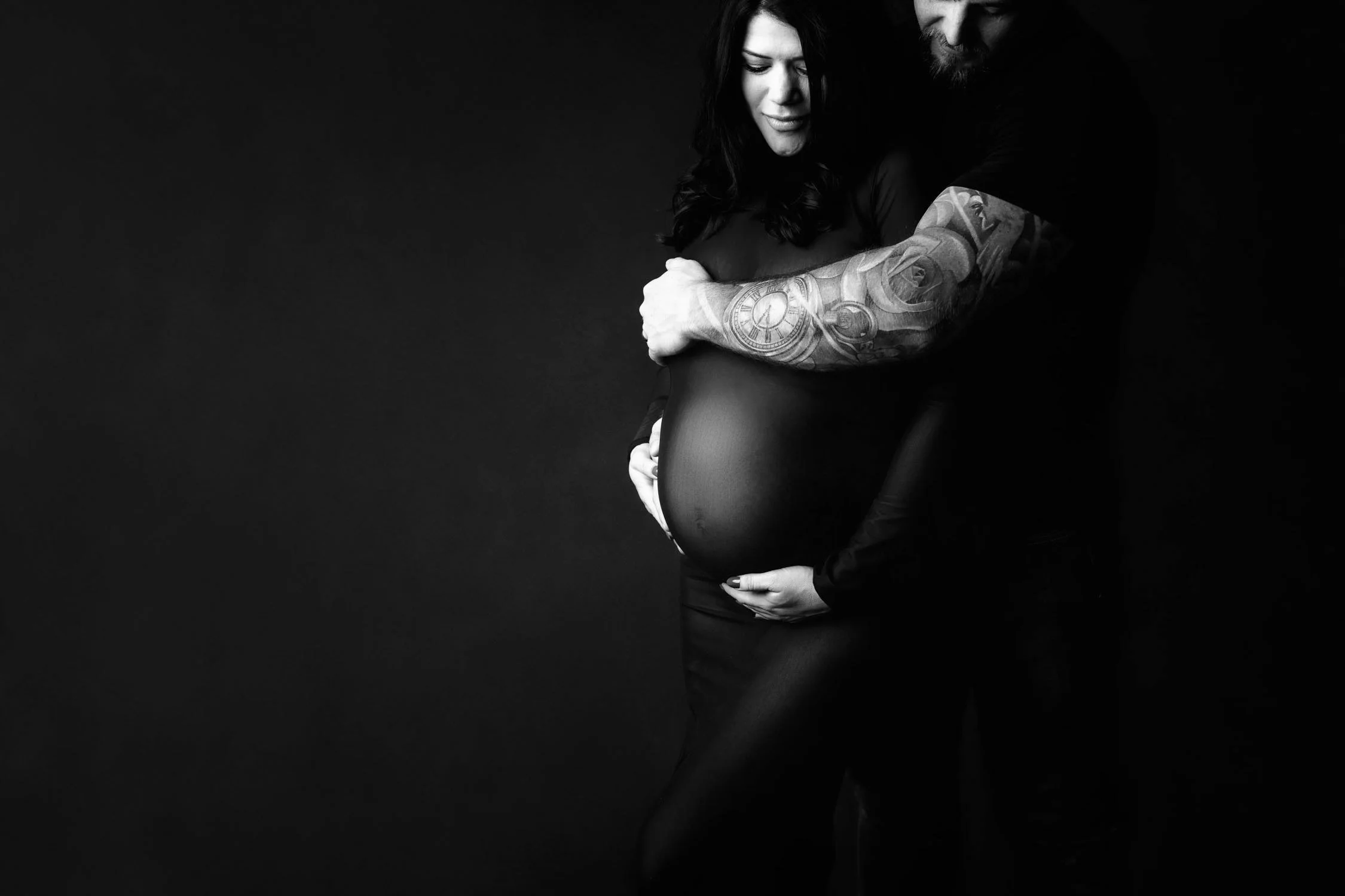 Black and white portrait of an expecting couple embracing with their hands around the mother’s baby bump.