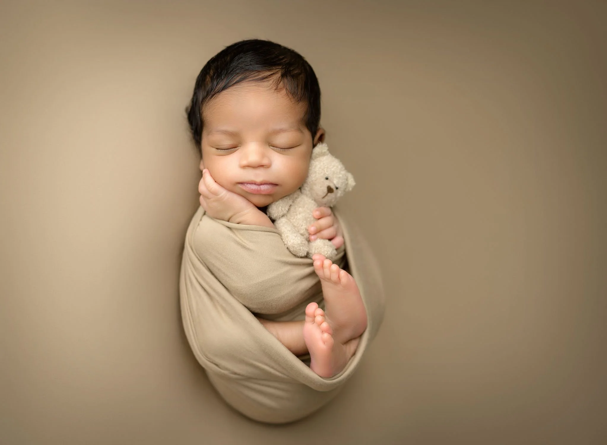 A newborn baby sleeping on its back in a tan swaddle and feet poking out with a white tiny bear