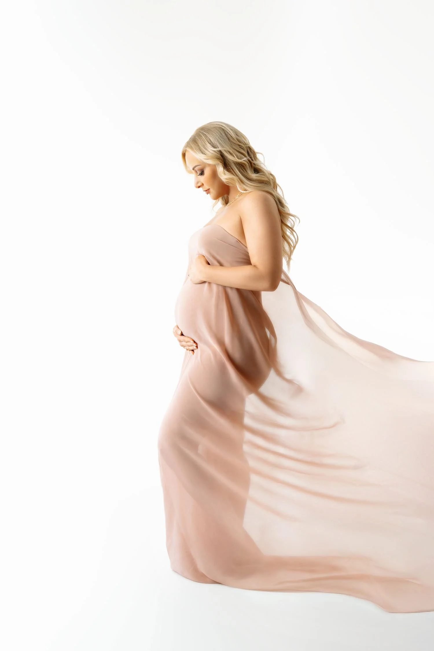 Pregnant woman in a flowing blush maternity gown holding her baby bump during a bright studio maternity portrait.
