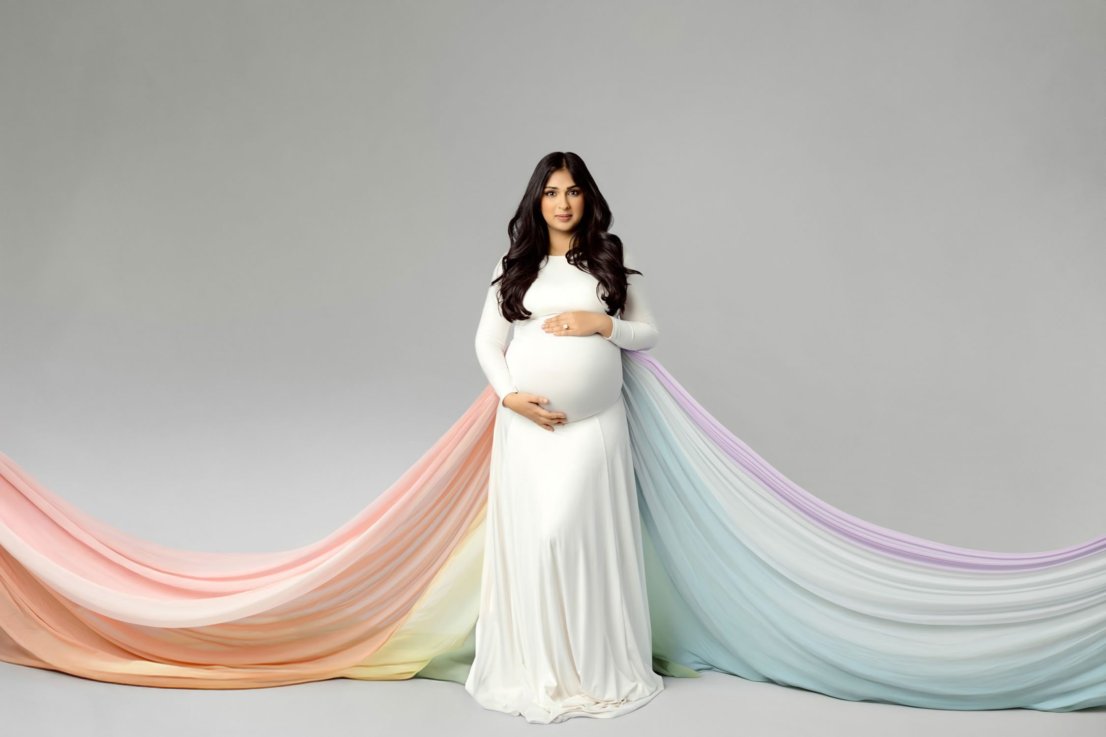 Expecting mother in a white gown standing in front of flowing pastel rainbow fabric during a St. Louis maternity session.