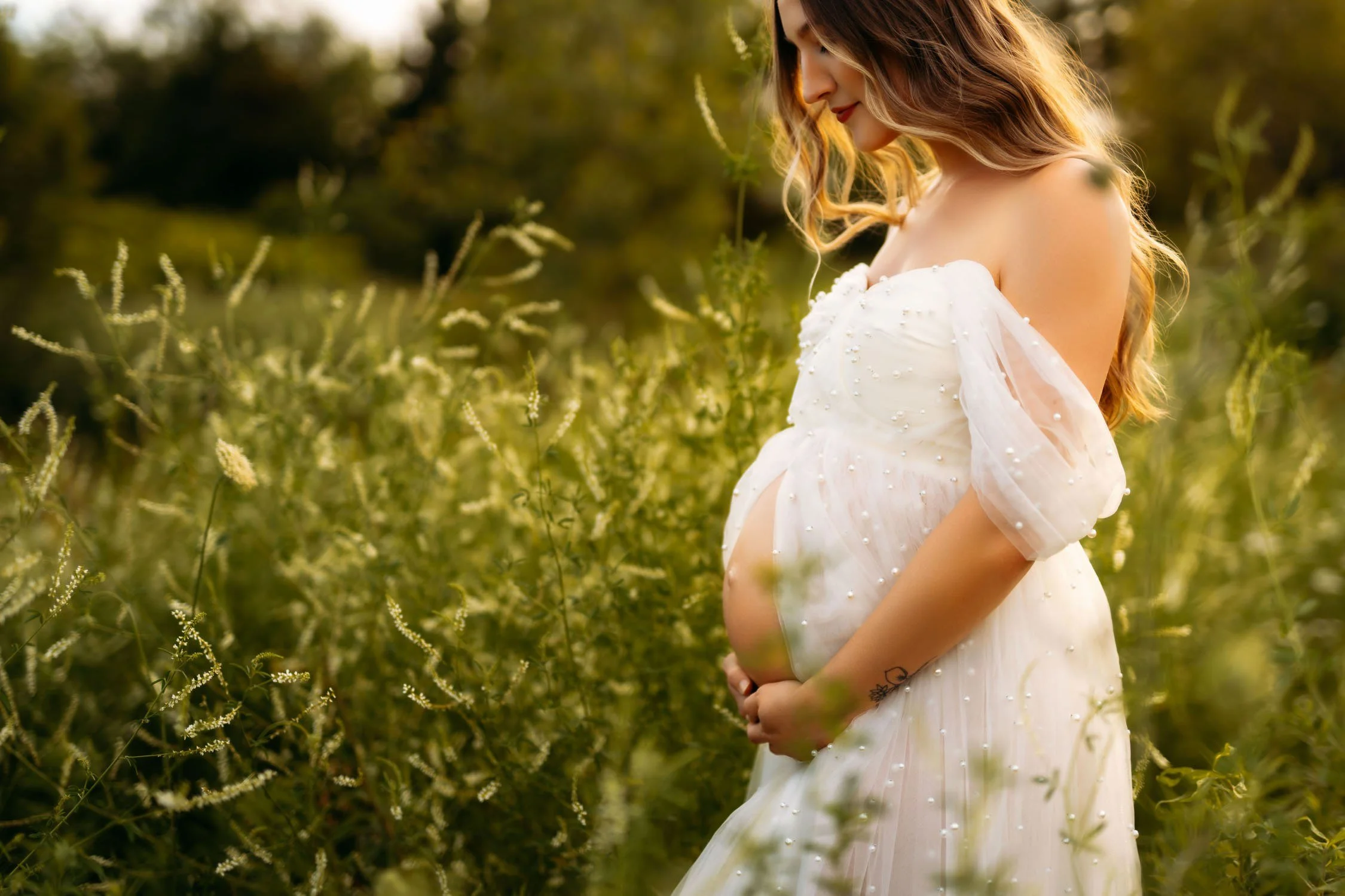 Pregnant woman in a flowing white dress standing among tall green grass during a St. Louis maternity session.
