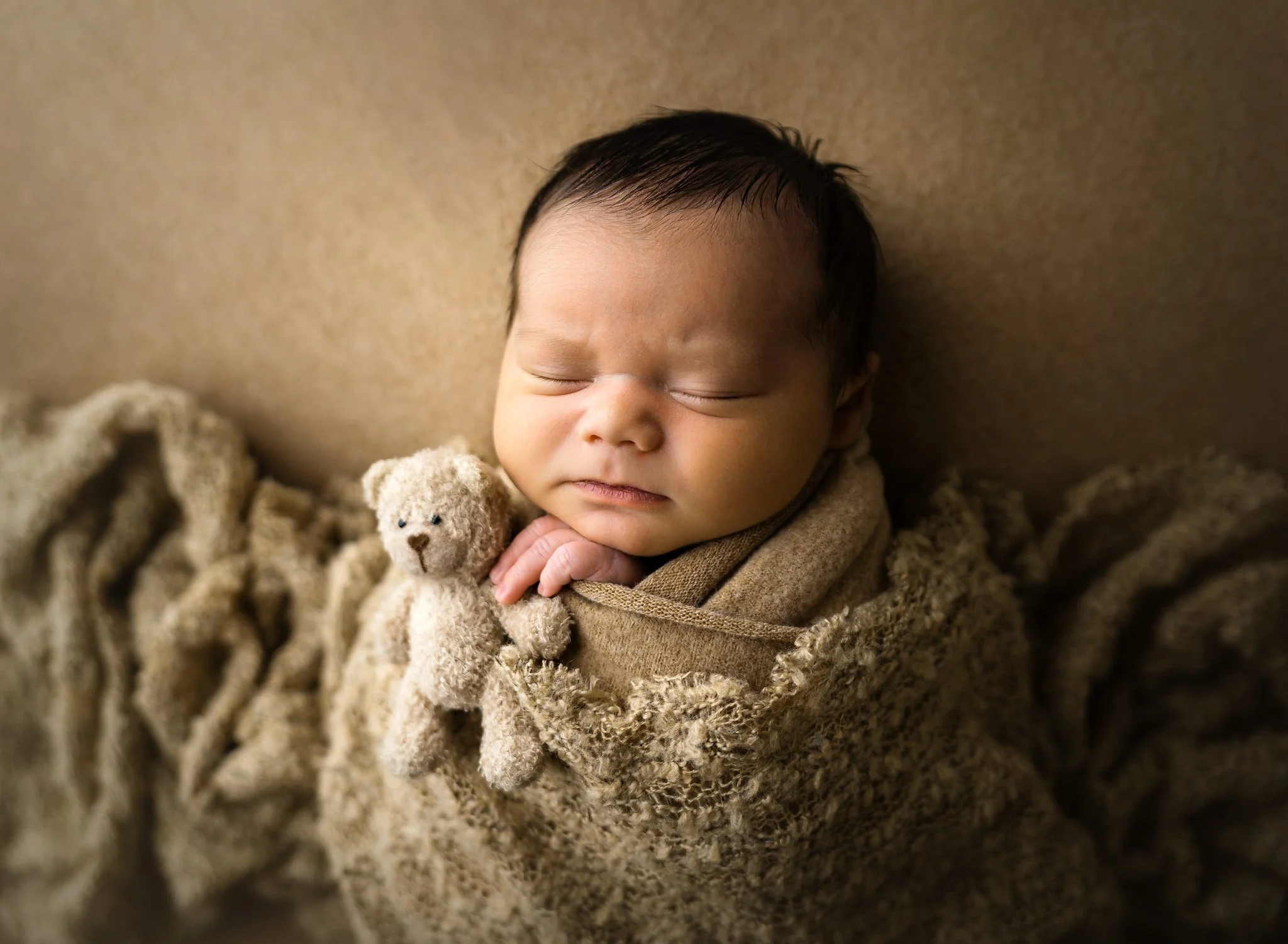A newborn baby sleeps holding a tiny bear under matching tan swaddle and blanket after meeting lactation consultants in st louis